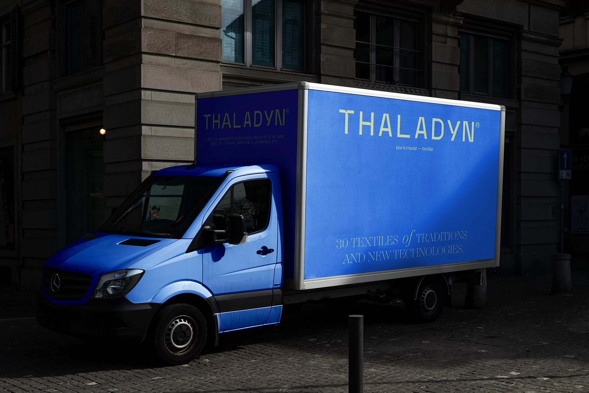 Blue delivery truck with 'THALADYN' branding parked on a cobblestone street, with urban building facade in the background, partially shaded.