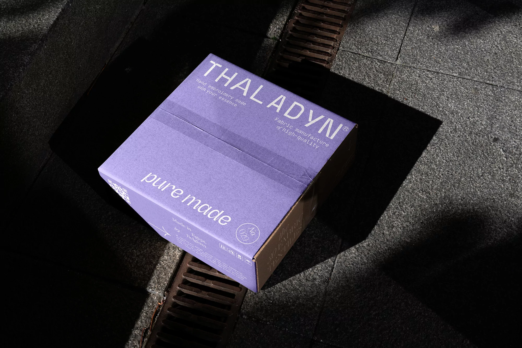 A lilac-colored box with product text sits on a stone surface with a grate casting a strong shadow across the scene.