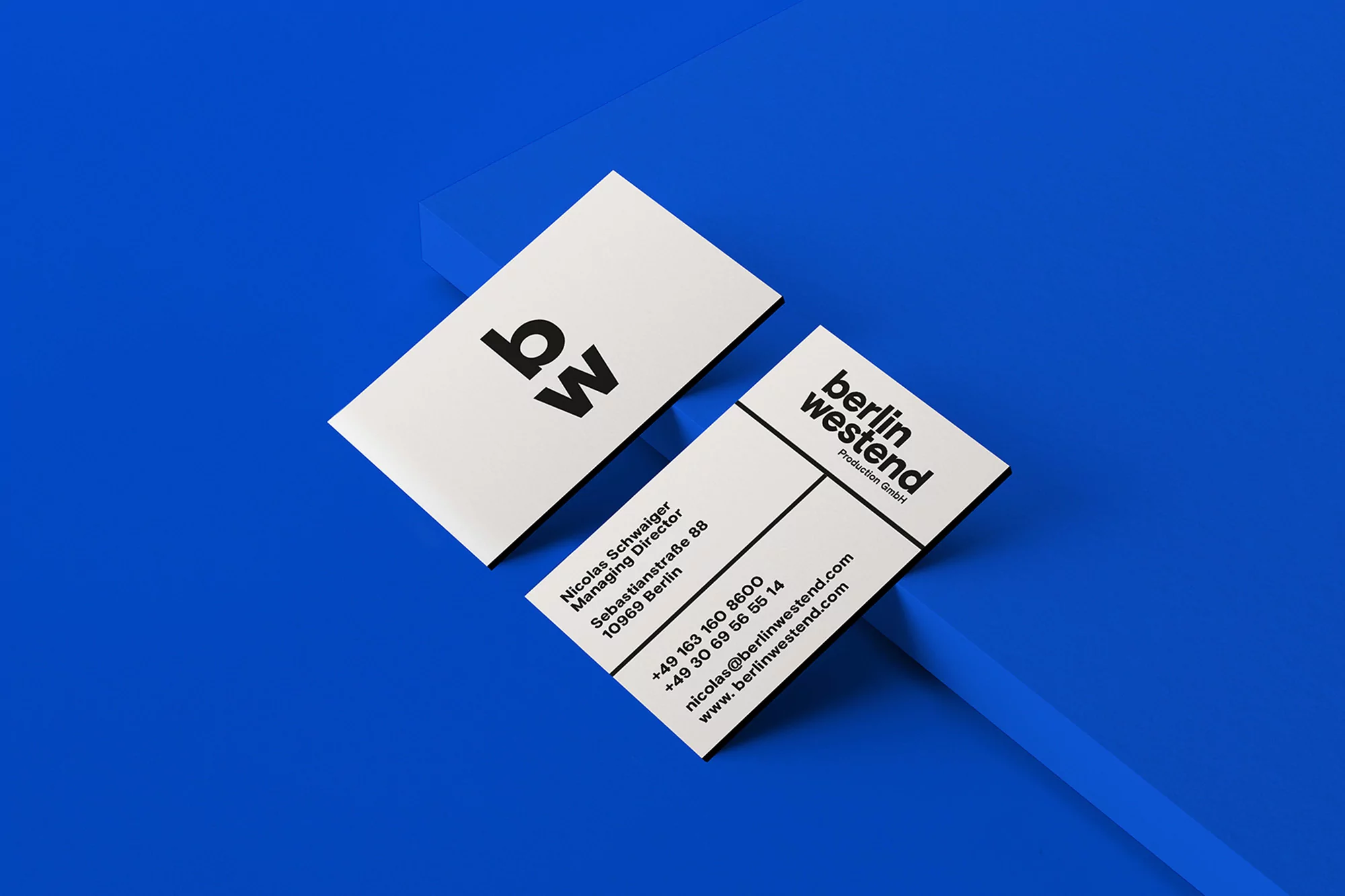 Two white Berlin Westend Production business cards with black text are angled on a solid blue surface, featuring company logo.