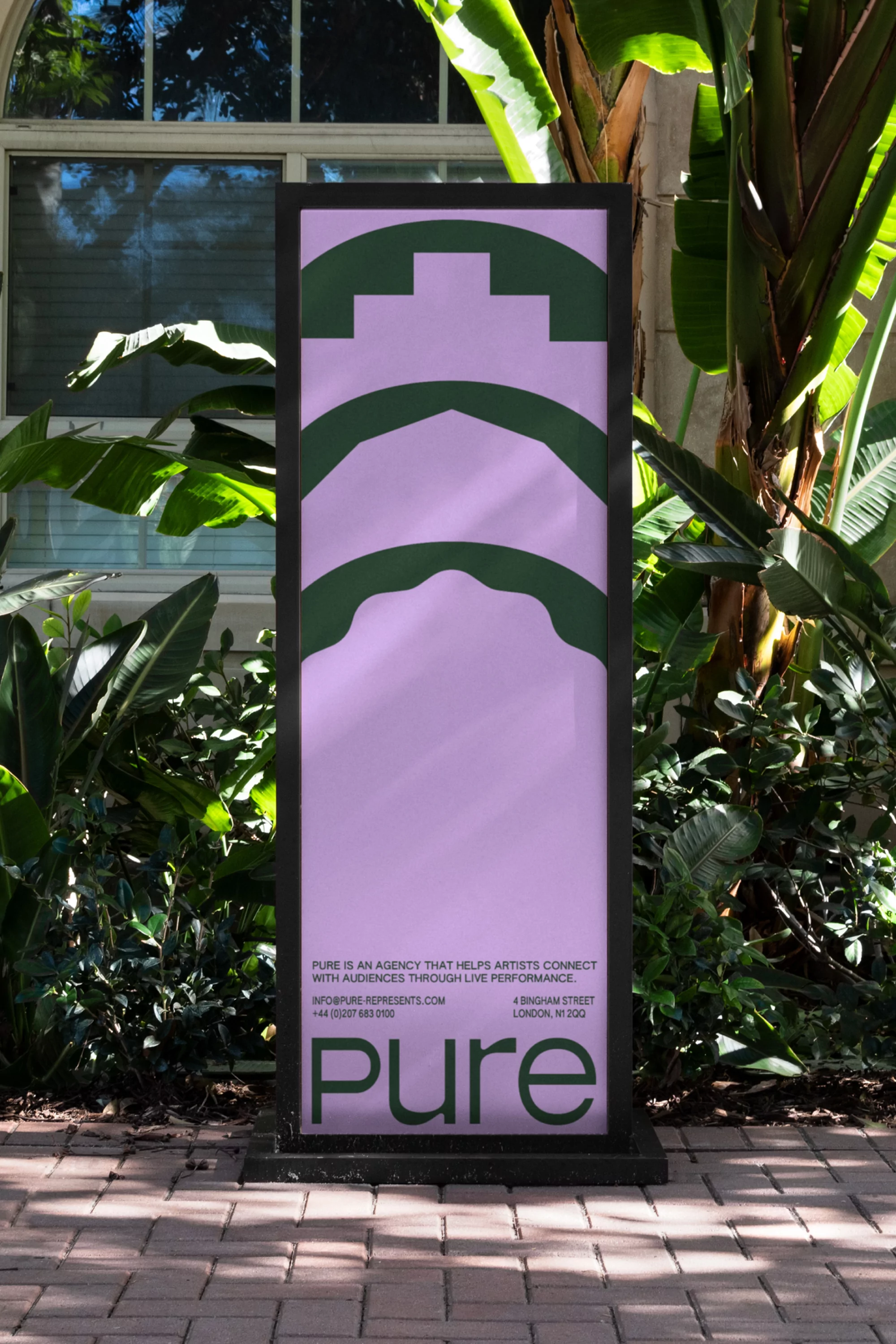 Rectangular sign featuring a logo and text, standing on a brick path with lush green foliage in the background, promoting a creative agency.