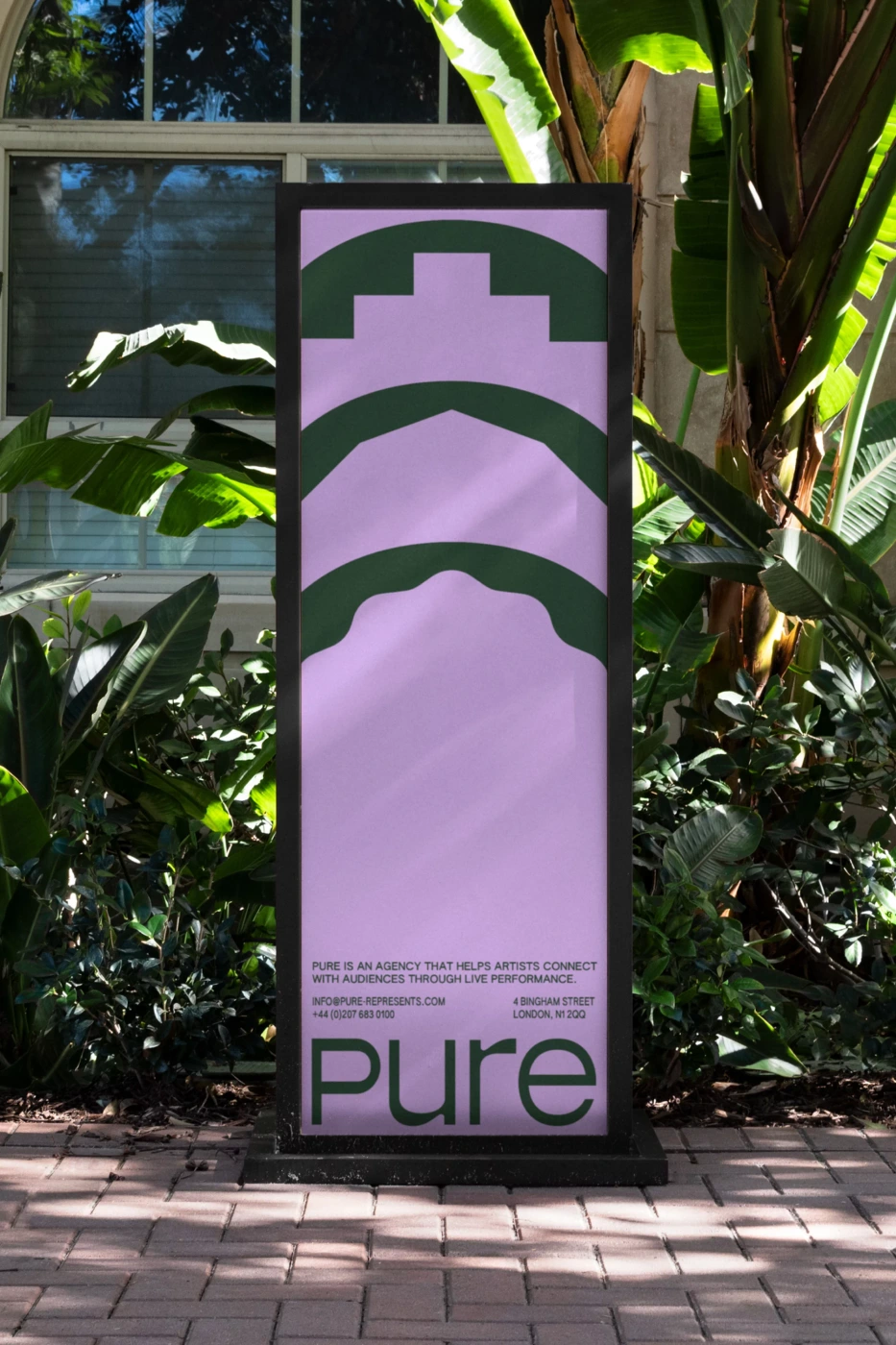 Tall branded sign with pink background and green geometric graphics on a paved path flanked by tropical foliage