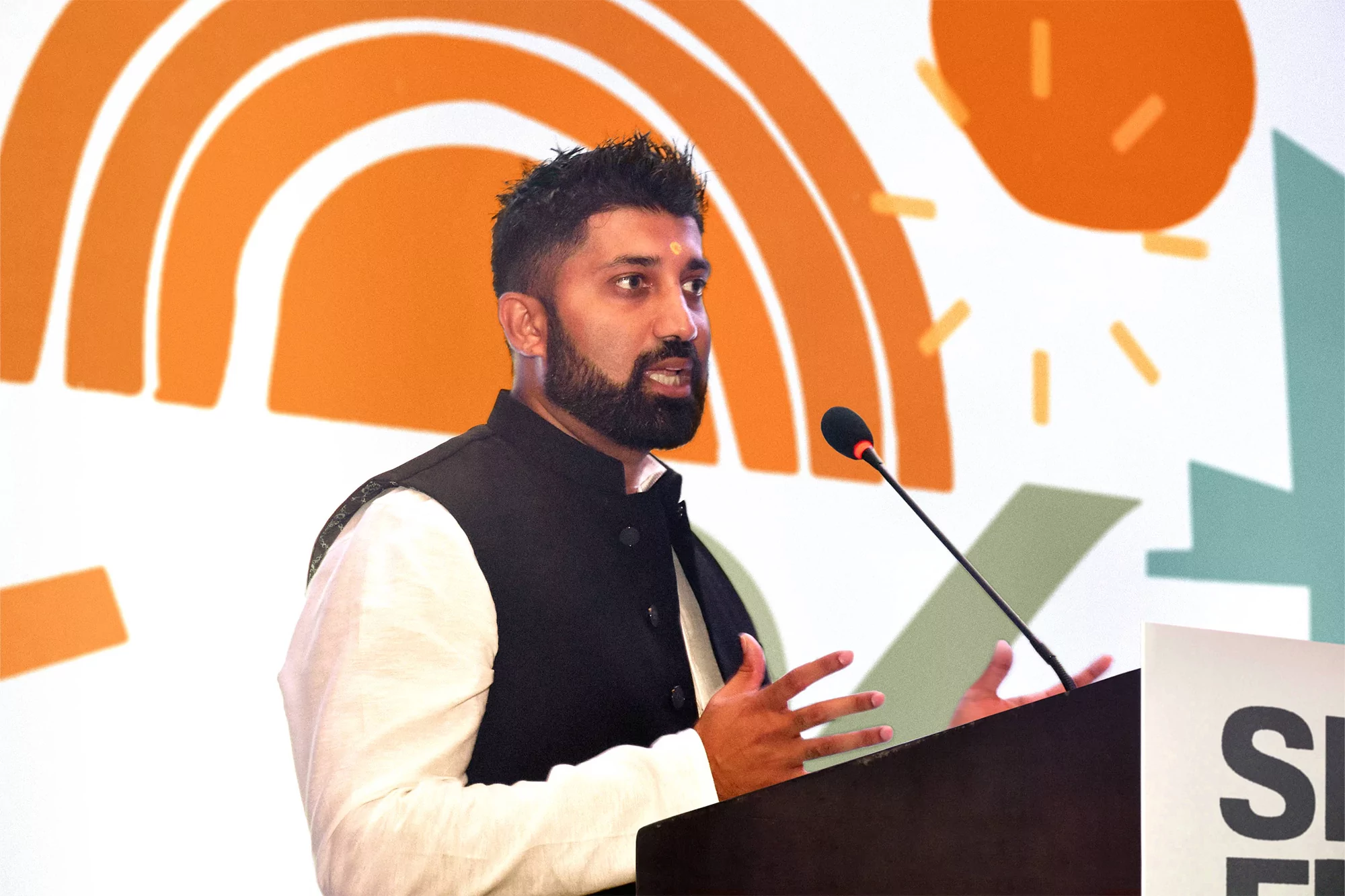 Adult man with beard presents at a podium in black vest over white shirt with orange graphic design in the background.