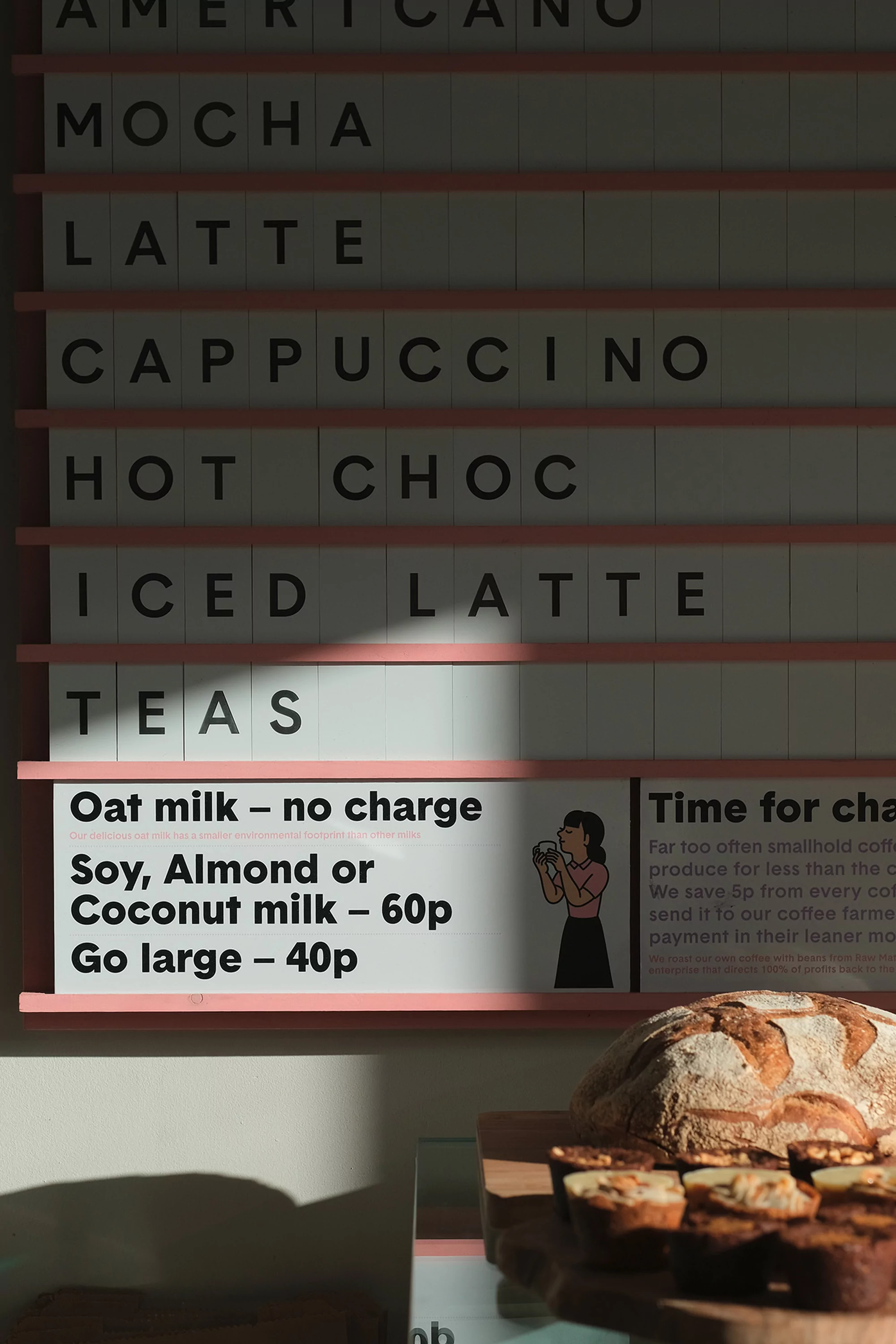 Cafe menu board with coffee and tea options, alongside bread on a counter. The scene is naturally lit creating a warm, inviting atmosphere.