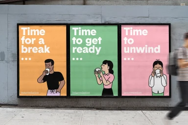 Three colorful promotional posters for Intermission mounted on a grey concrete wall with a blurred pedestrian walking past
