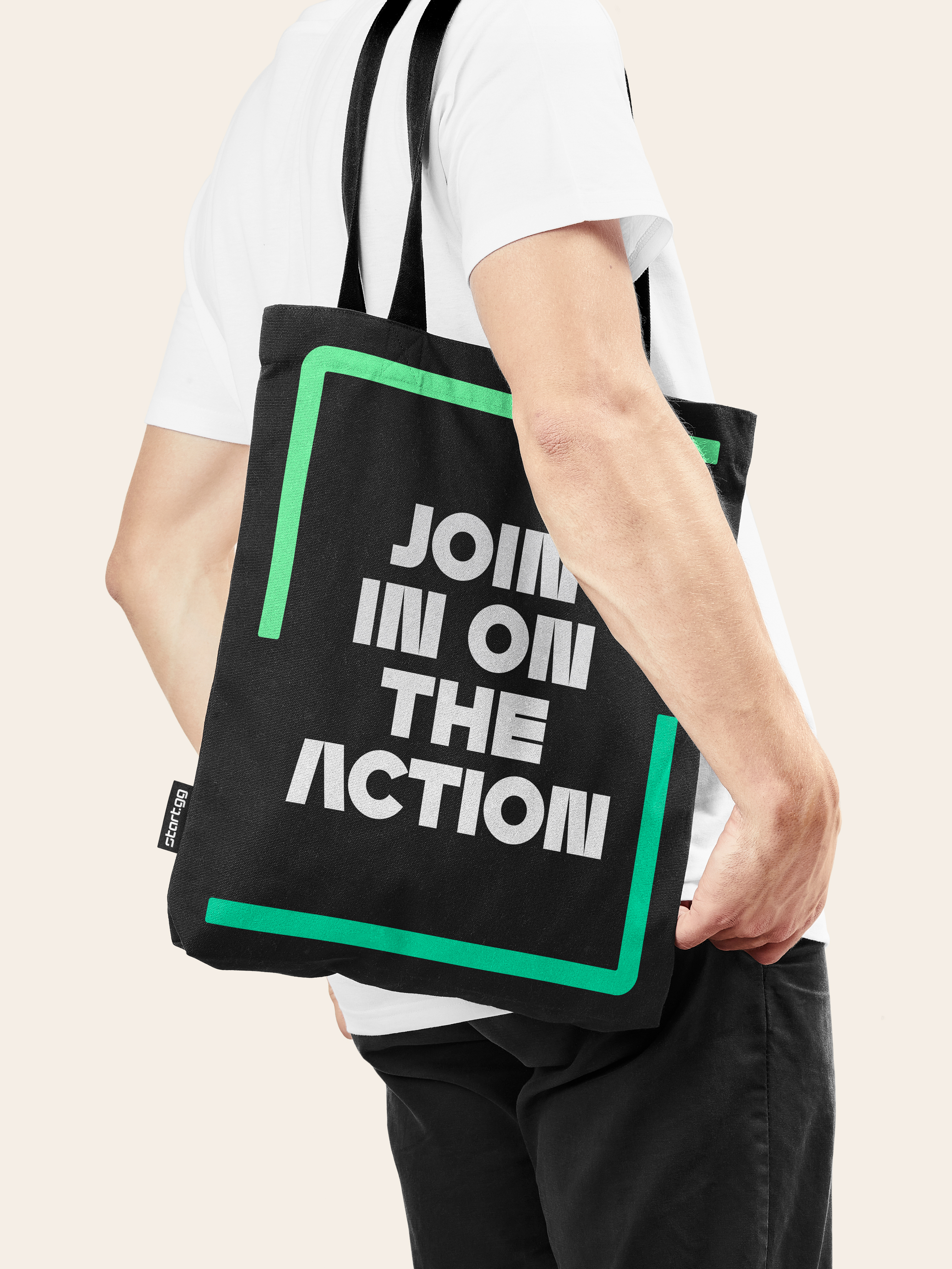 Adult man wearing a white t-shirt carrying a black tote bag with white text and a green border against a pale pink background.