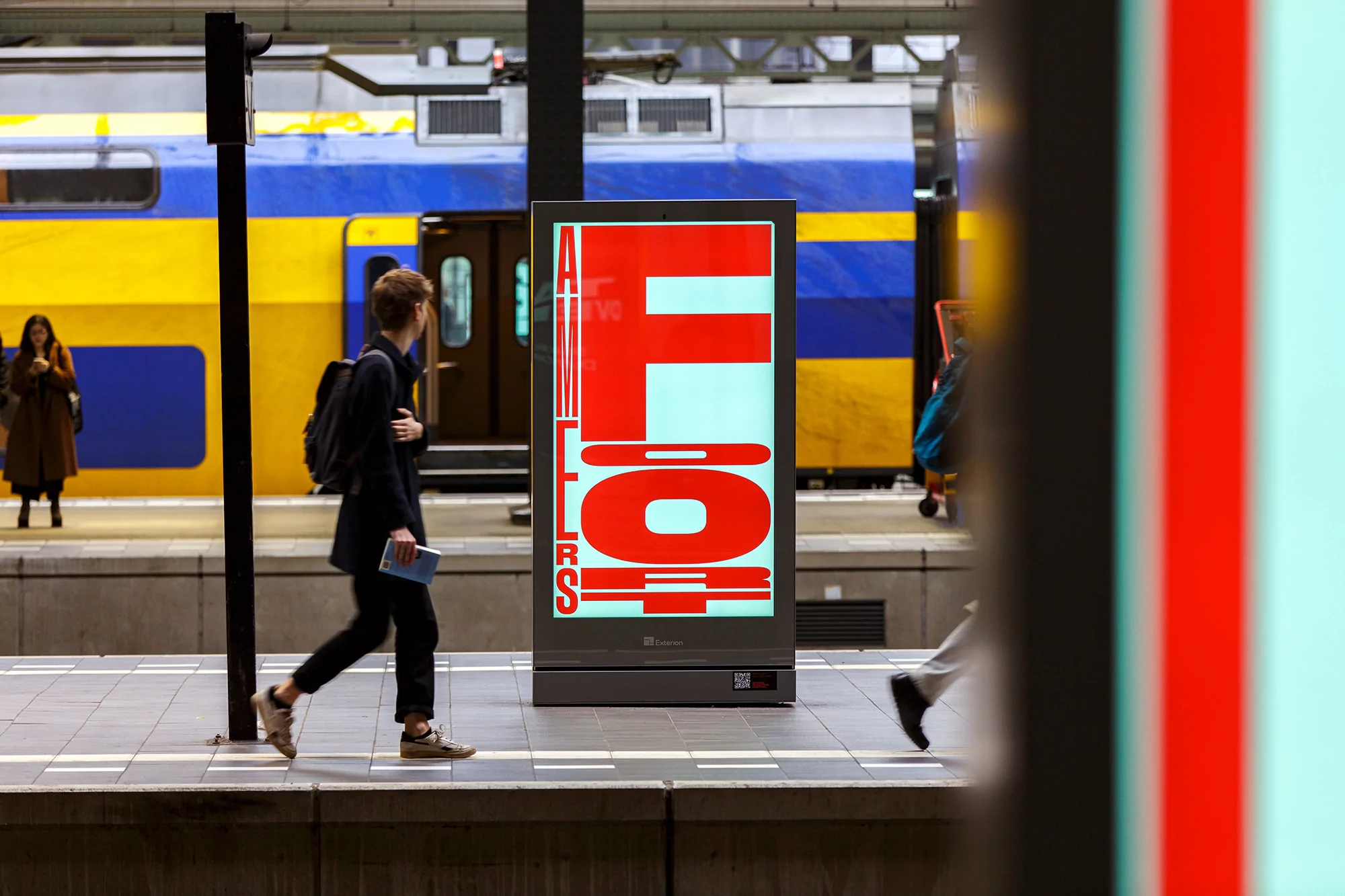 Illuminated advert at a train station with a train in the background and a man walking past.