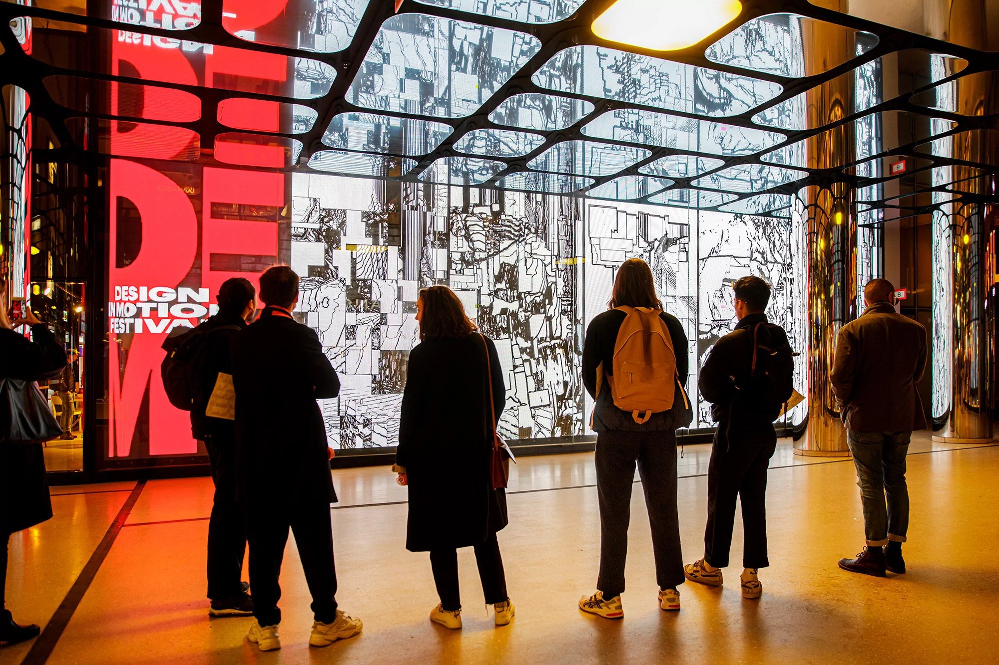 A group of people stand in a modern building, gazing at a large screen displaying red text and black and white digital art.