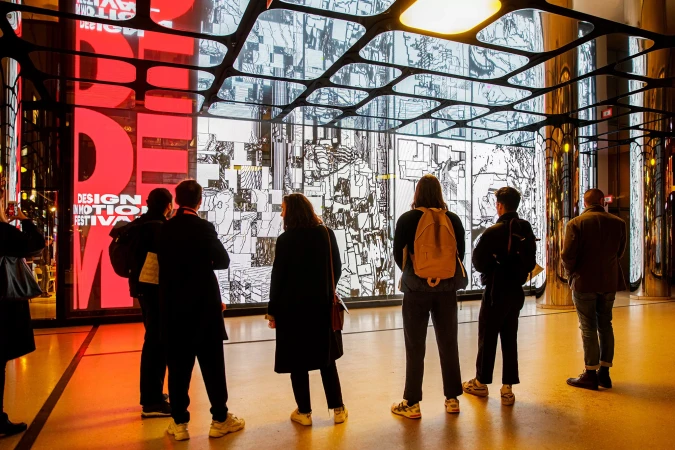 A group of people stand in a modern building, gazing at a large screen displaying red text and black and white digital art.