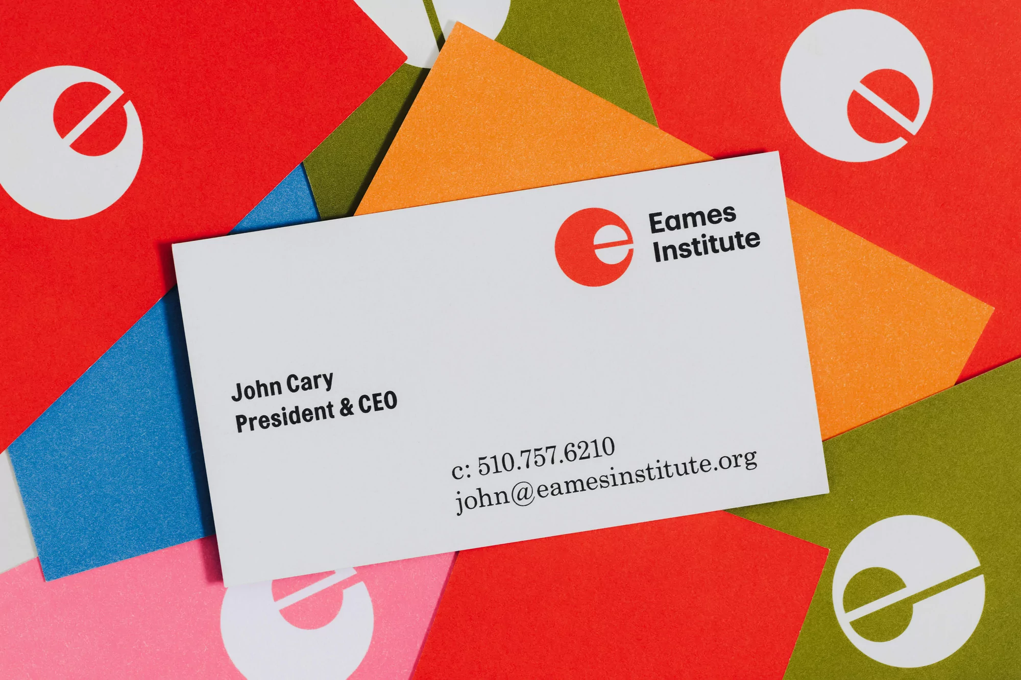 Close-up of a white business card for John Cary, President & CEO of Eames Institute, resting on colorful paper with logo patterns.