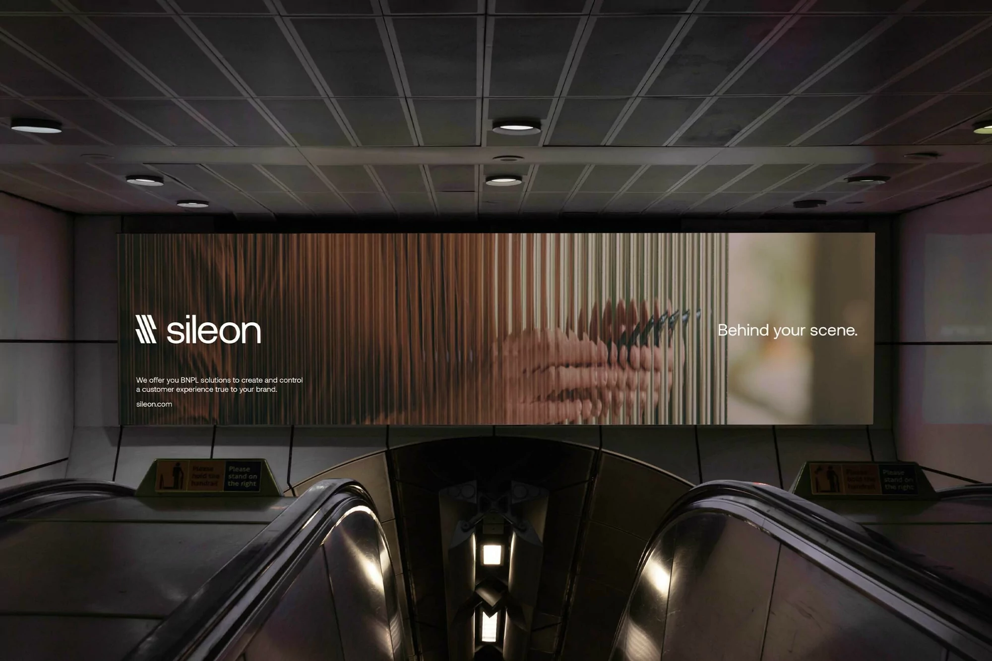 Illuminated Sileon brand advertisement in a subway station over an escalator, featuring abstract depiction of hand holding phone.
