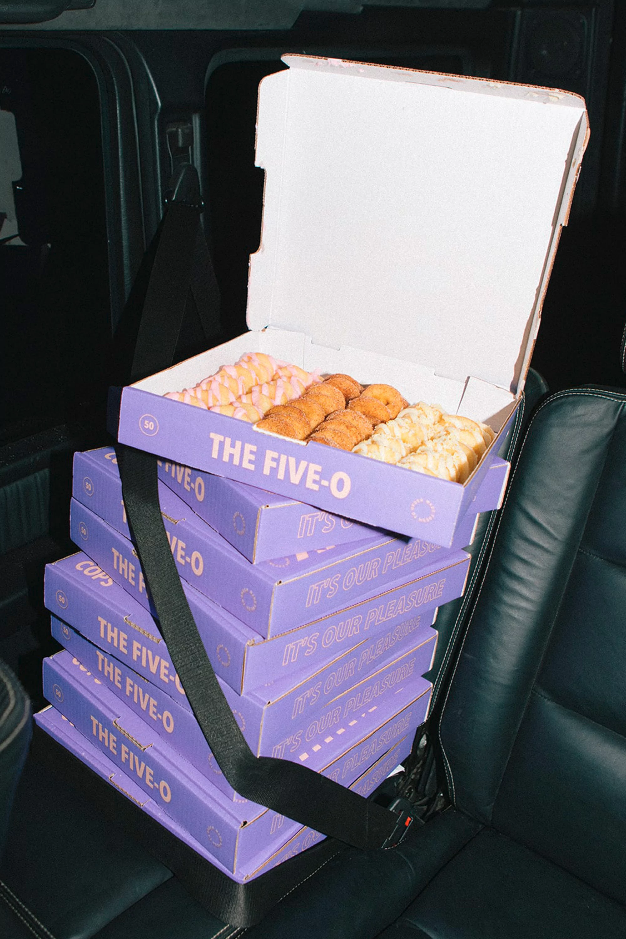 Stack of purple donut boxes from The Five-O displayed on a black leather car seat, one box open, revealing an assortment of donuts.