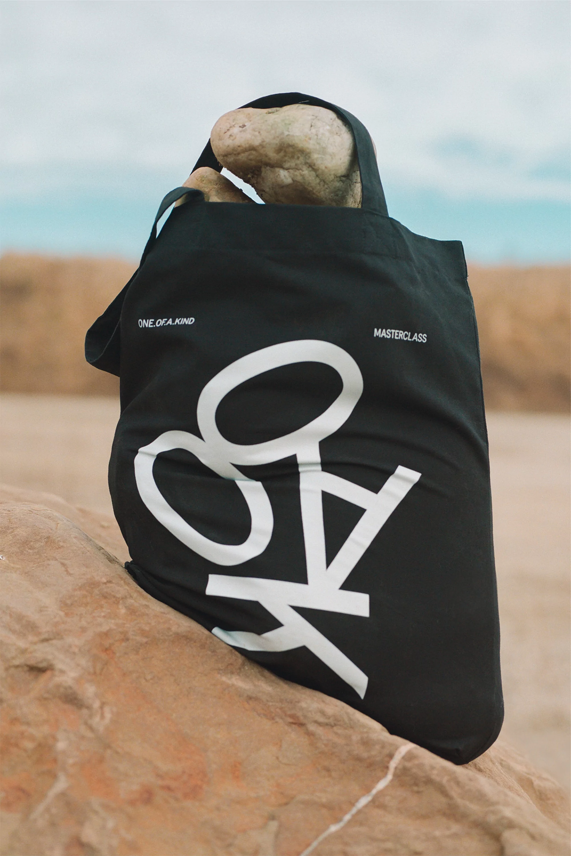 Black tote bag with white graphic print on a tan-colored rock outdoors. Rocks fill the bag on a cloudy day near the ocean.