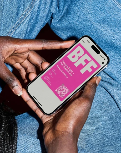 Close-up of a person holding a smartphone displaying a digital ticket with 'BFF' in pink, against a denim-clad background.