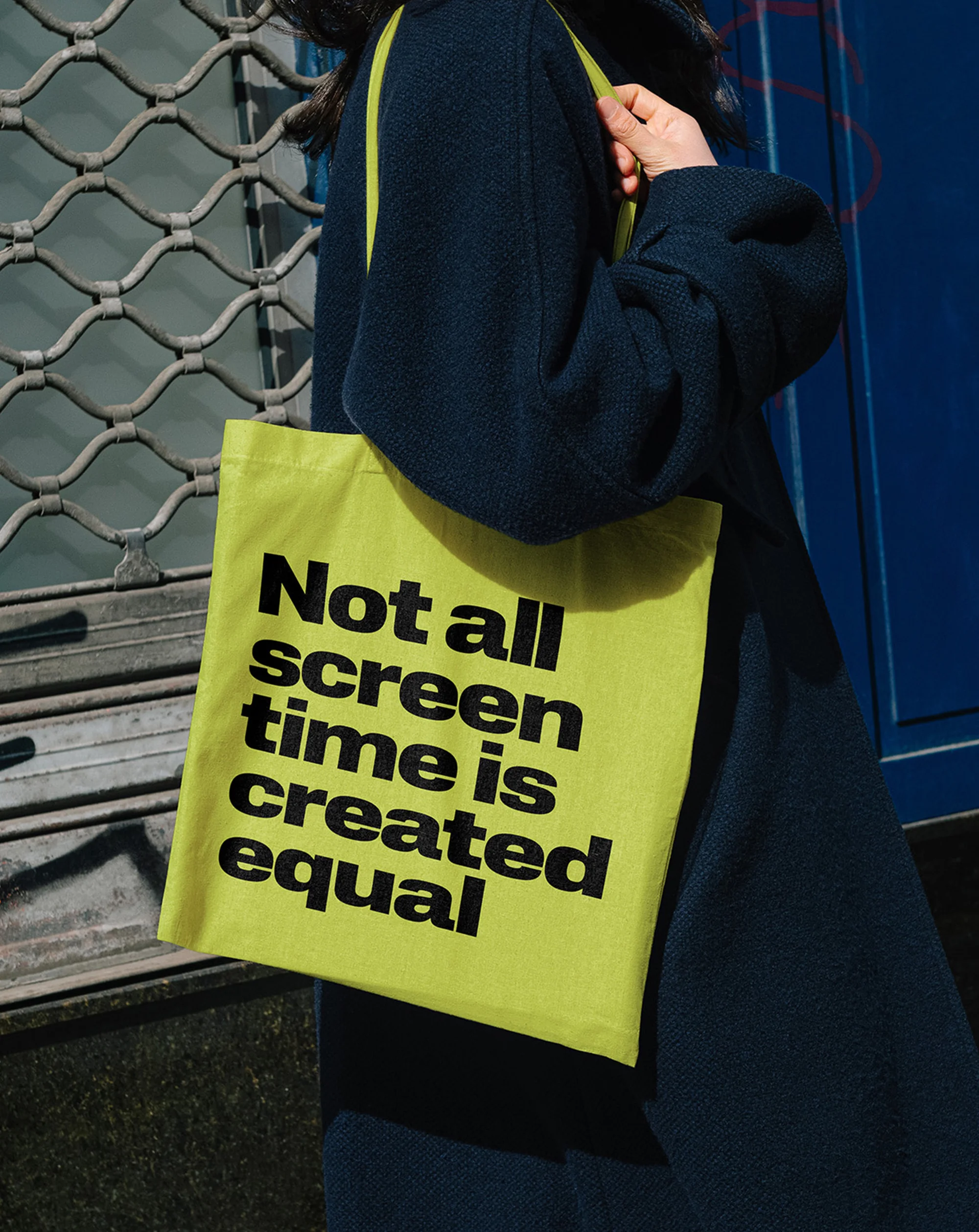 Person in a dark coat carries a bright yellow tote bag with the black text: 'Not all screen time is created equal.'