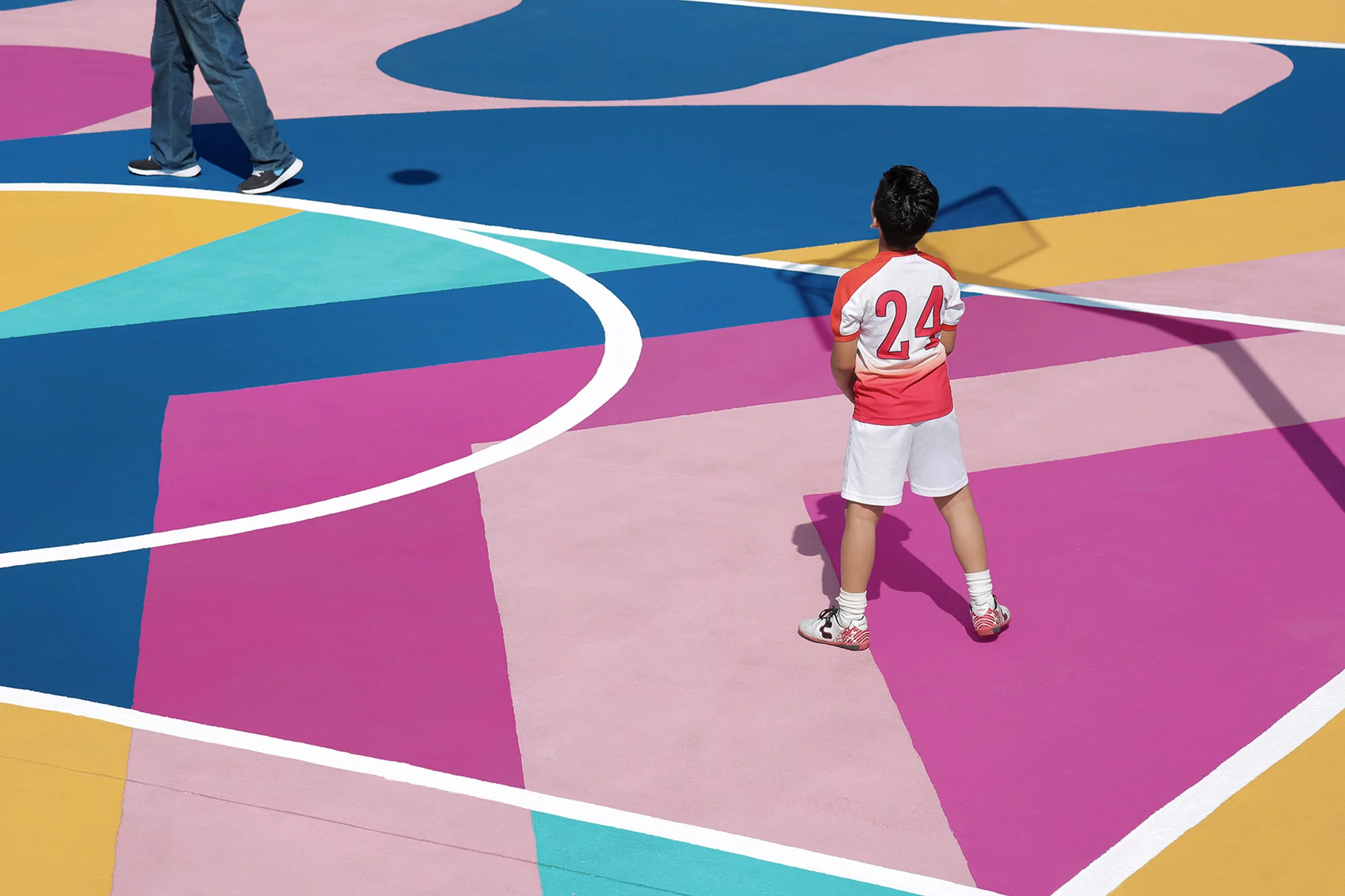 A child wearing a number 24 jersey stands on a multicolored, geometric patterned outdoor court with a partial view of another person's legs.