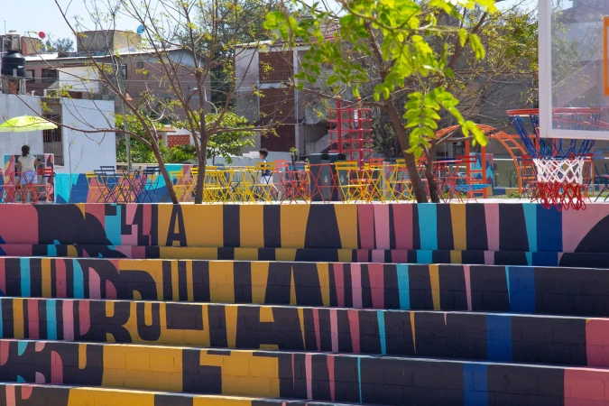 Outdoor gathering space with colorful painted stairs, metal furniture, greenery, and a basketball hoop in a sunny urban setting.