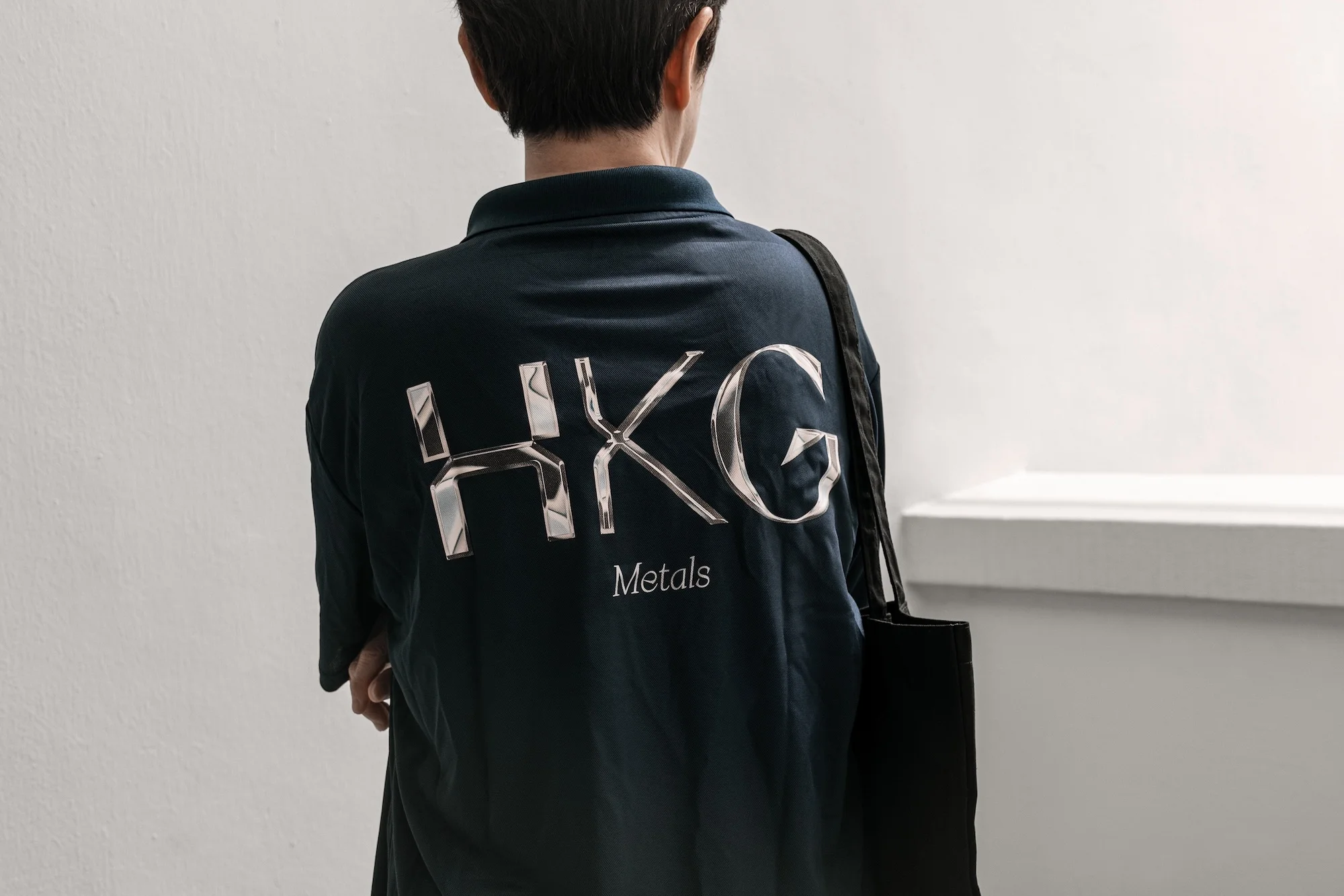 Back view of a person wearing a dark polo shirt with 'HKG Metals' in reflective silver, paired with a black tote bag indoors.