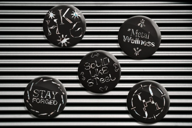 Five black pinback buttons with white metallic stylized text designs are displayed against a black and gray corrugated metal backdrop.