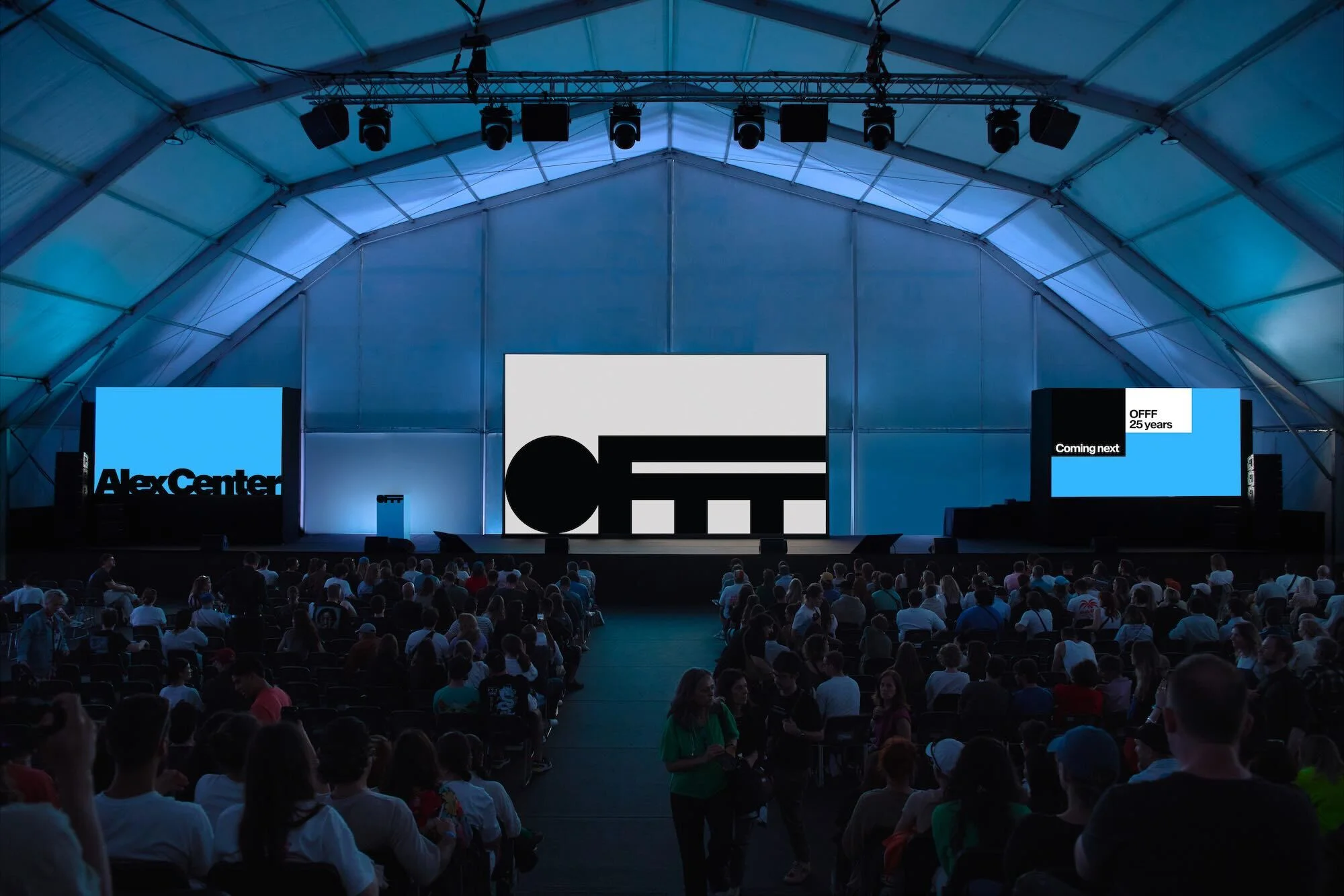 Large crowd attends a presentation on stage with LED screens at an event under a tent with blue lighting and a dark foreground.