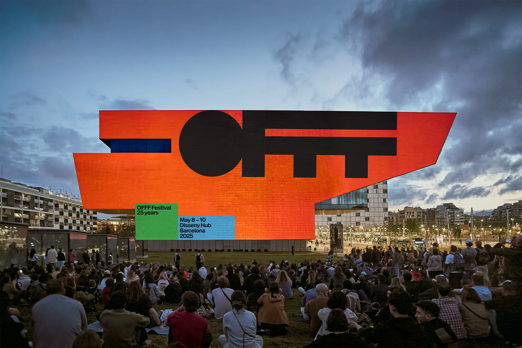 Large crowd of people sit on a lawn facing an orange building with black text at the OFFF Festival in Barcelona at dusk.