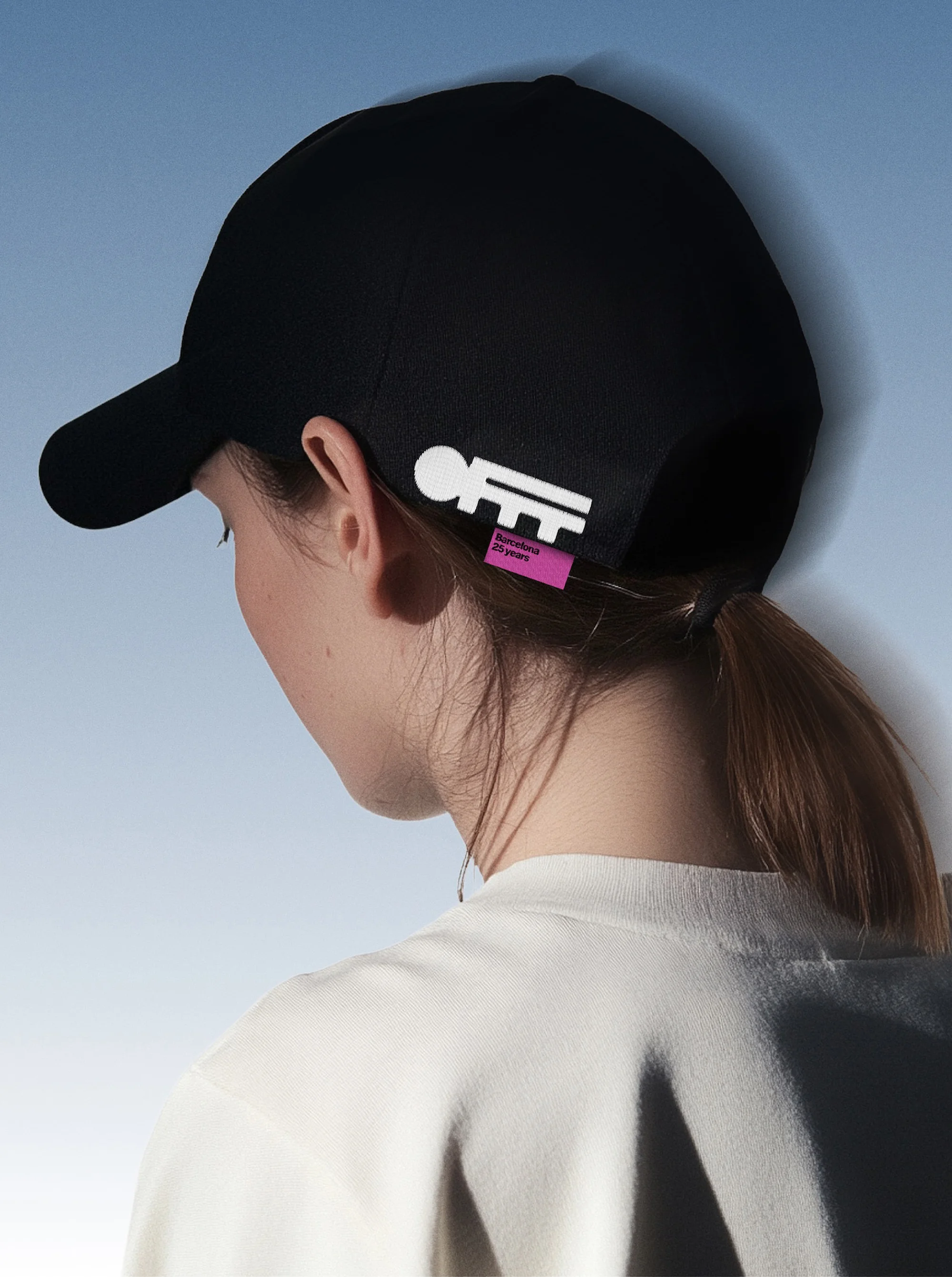 Young woman wearing black cap with white and purple logo, posing against a blue sky backdrop in a fashion forward outdoor setting.