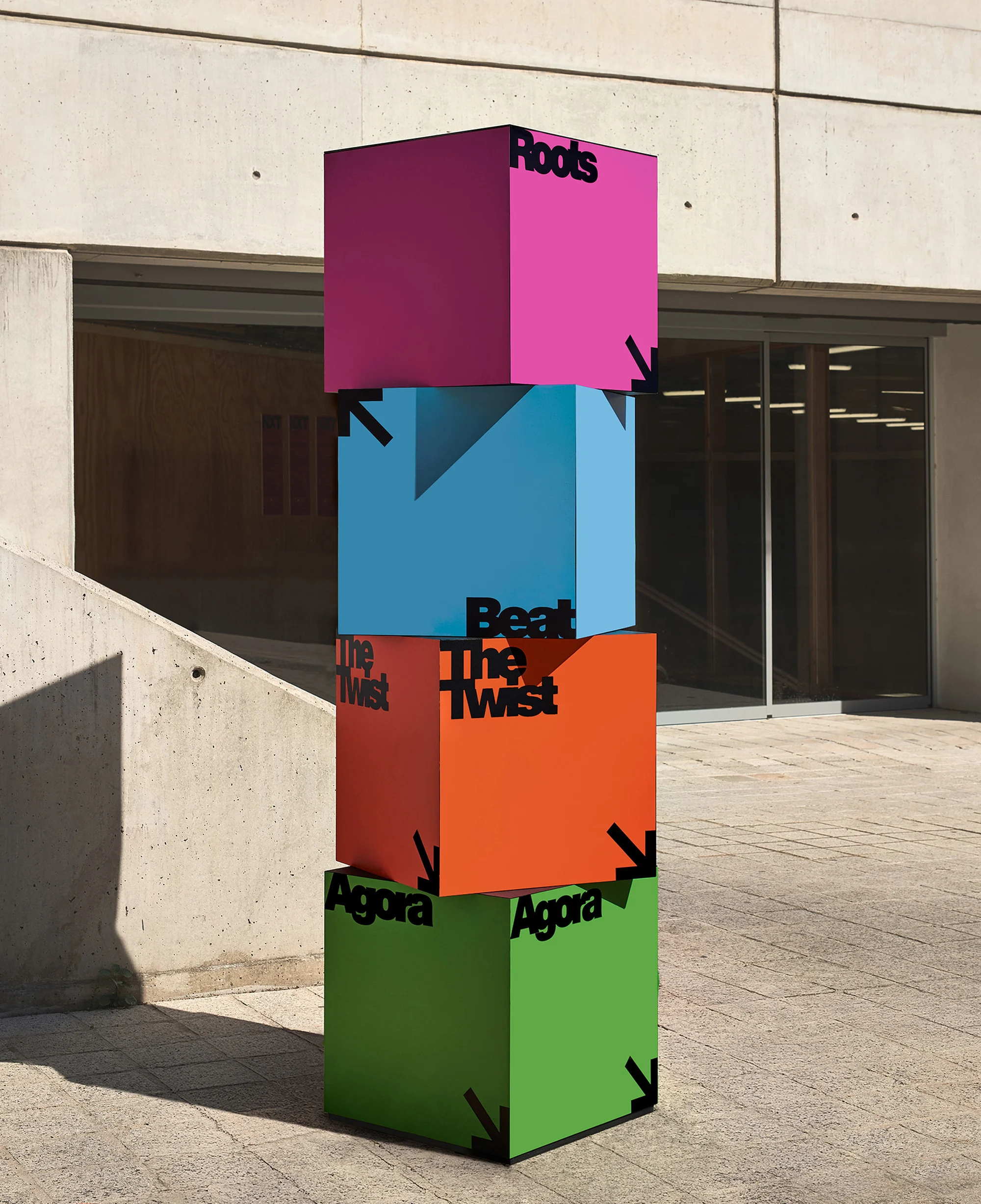 Colorful cubic sculpture featuring typography displayed outdoors. Stacked boxes in pink, blue, orange, and green with black text and arrows.