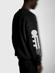 Profile view of a black man wearing a black sweatshirt with large white typographic print on the sleeve against a white wall