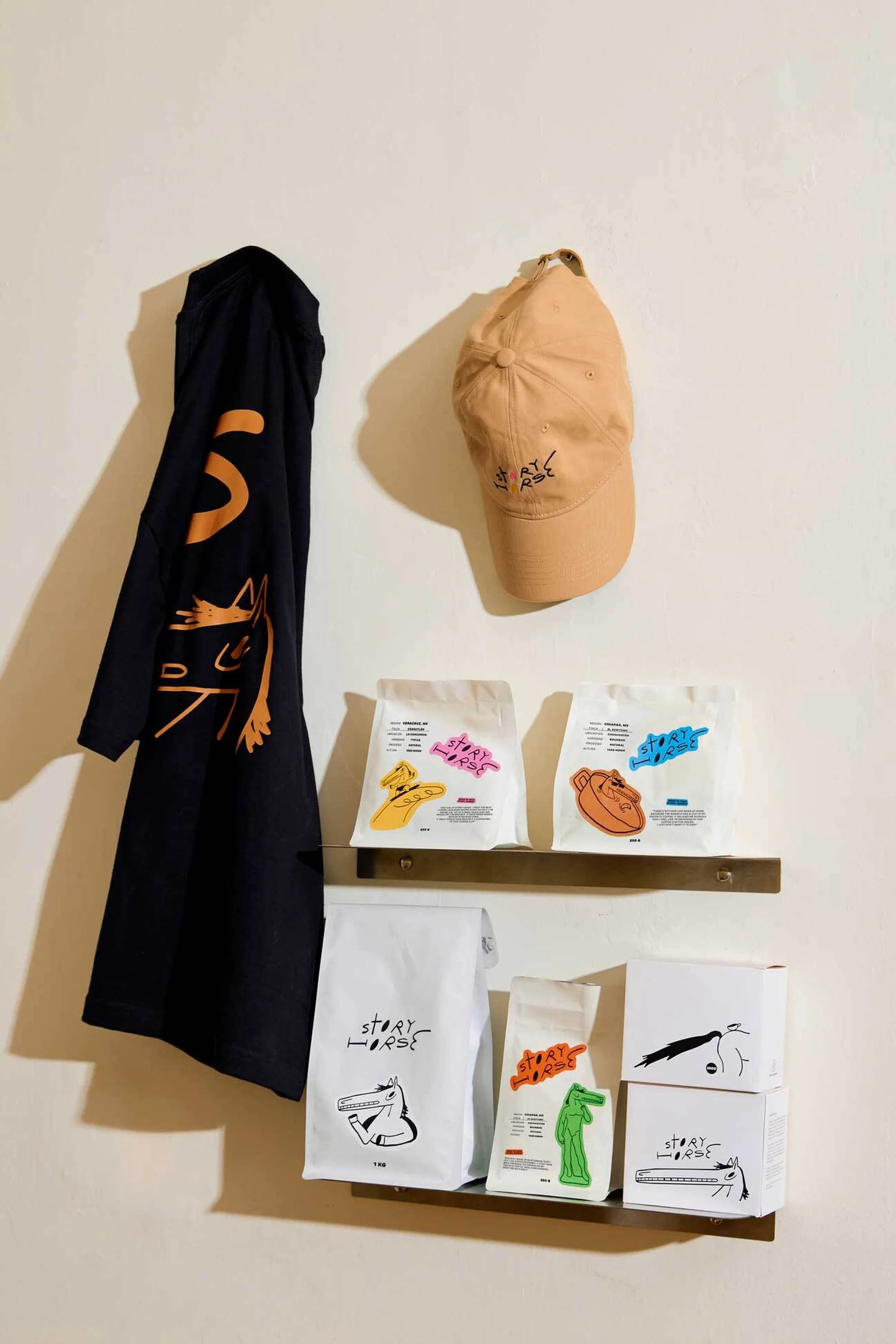 Still life showcases black jacket, tan hat, and colorful bagged goods on shelves against cream wall, creating playful, curated display.