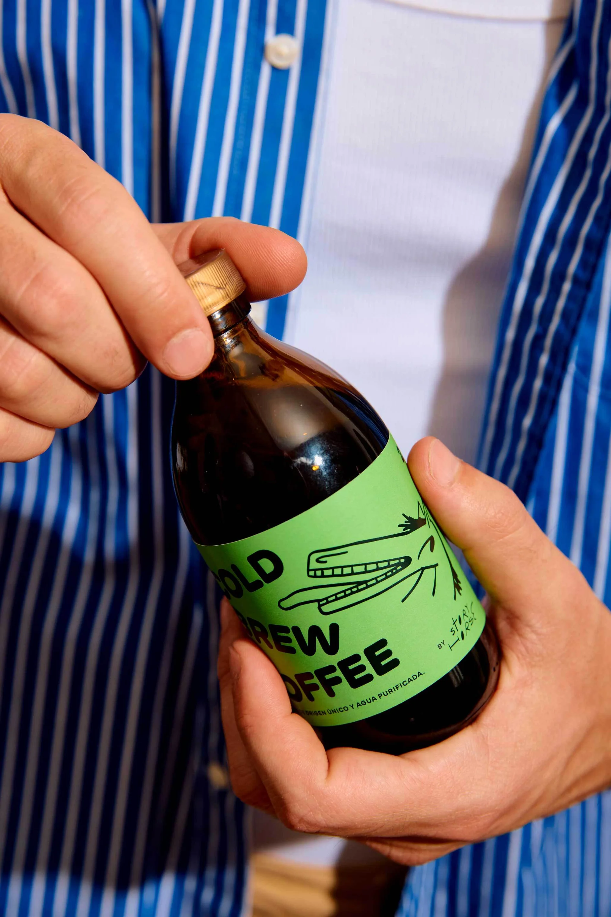 Person holding a bottle of cold brew coffee with a light green label with a cartoon alligator with a blue and white striped shirt.