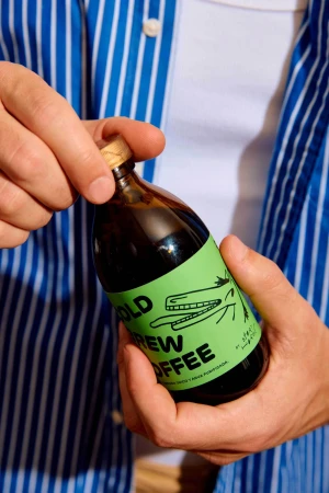 Person holding a bottle of cold brew coffee with a light green label with a cartoon alligator with a blue and white striped shirt.