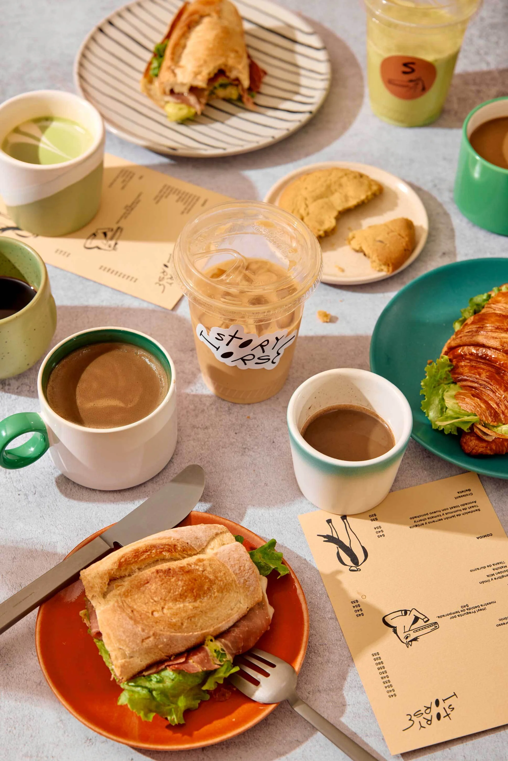 Overhead shot of table with sandwiches, croissant, coffee drinks, and cookie on plates. Food photography displaying cafe menu items.