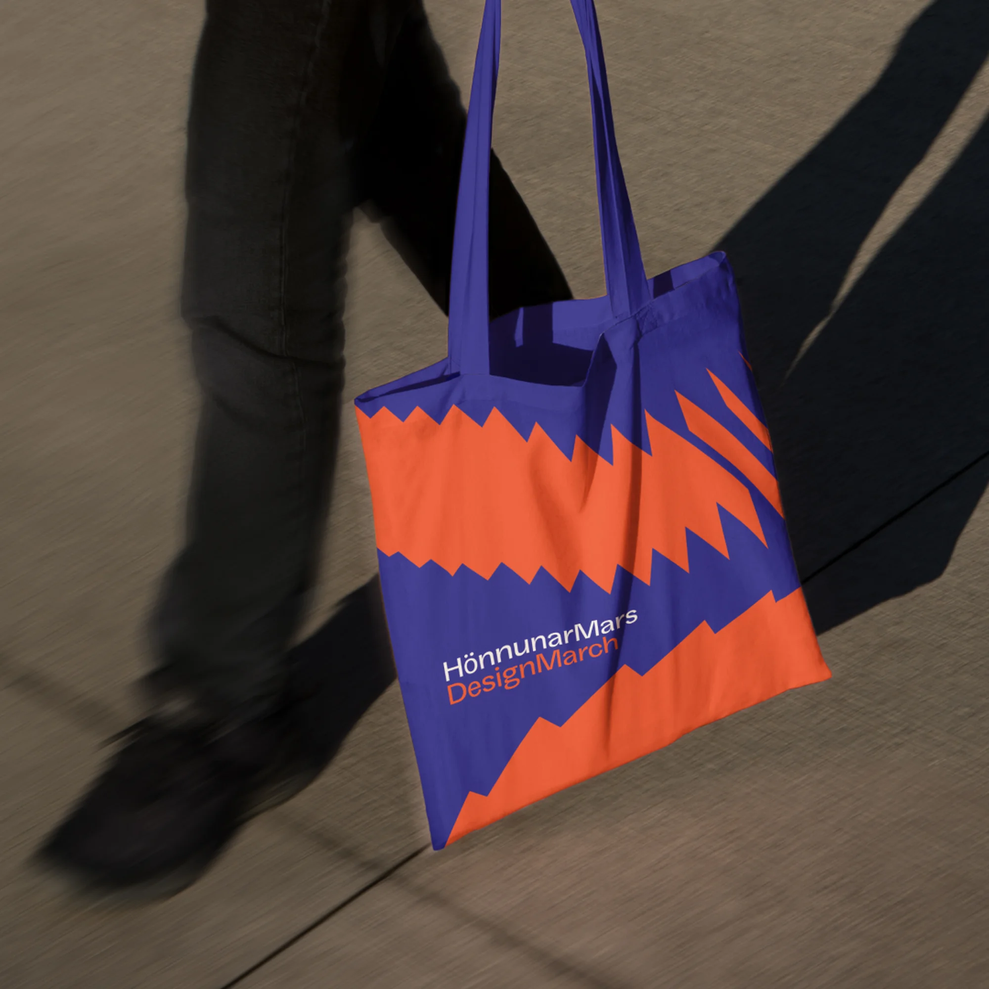 A blue and orange tote bag hanging on a shoulder with the words "HönnunarMars DesignMarch" printed on the bottom left of the bag.