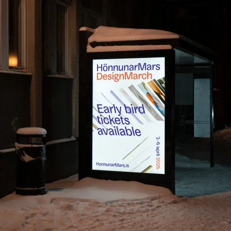 Illuminated advertising poster featuring 'HönnunarMars DesignMarch' visible at a bus stop in a snow-covered urban setting at night.