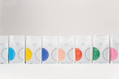 A line of white coffee bags with colourful circular labels and typography arranged on a seamless white studio background.
