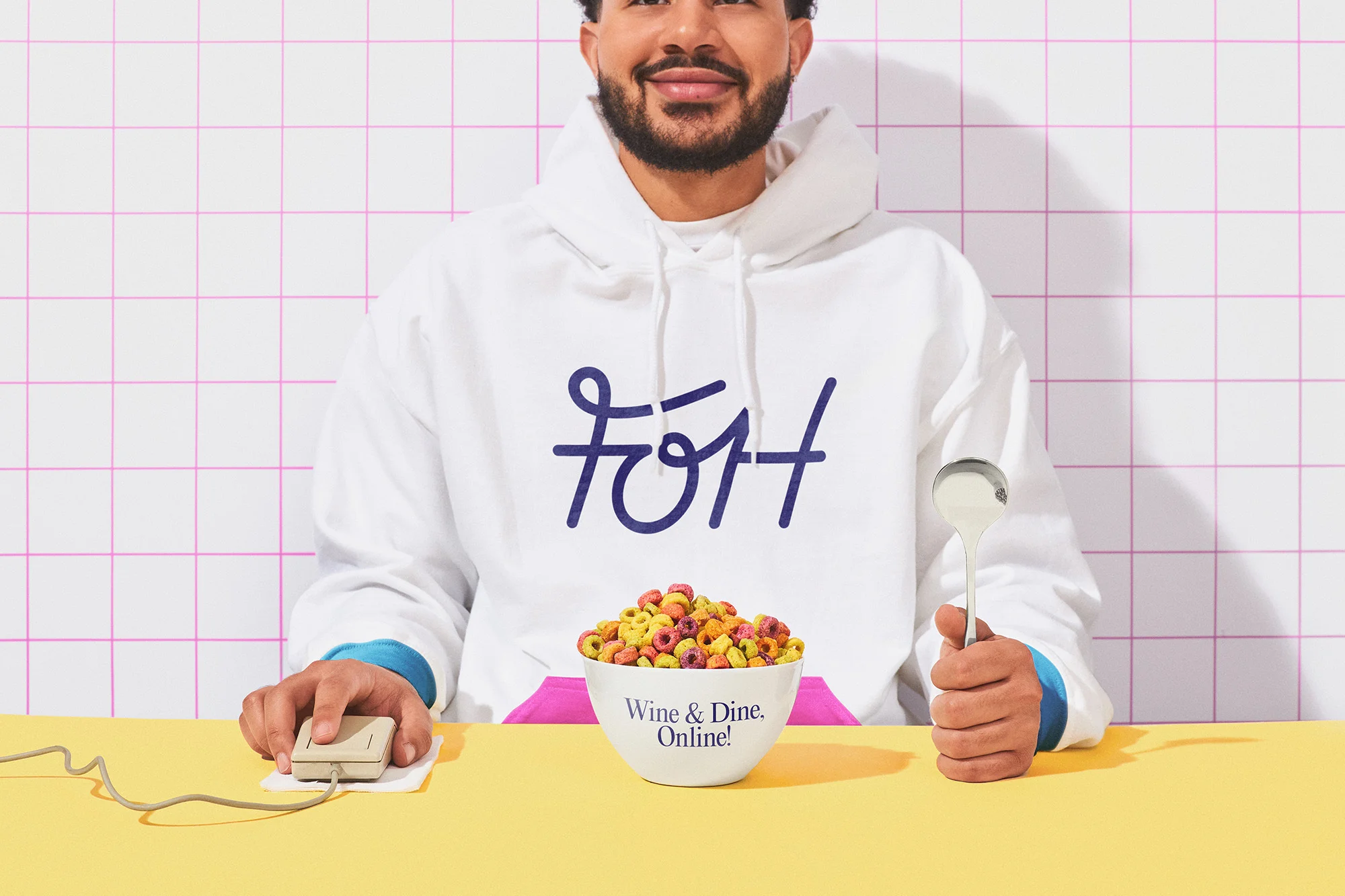 Person in a white hoodie holding a bowl of cereal and a spoon, with a retro computer mouse on a yellow table against a tiled wall.