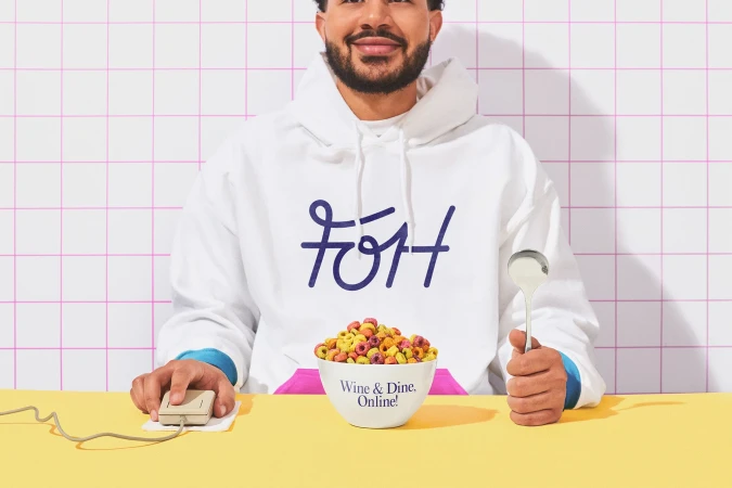 Person in a white hoodie holding a bowl of cereal and a spoon, with a retro computer mouse on a yellow table against a tiled wall.
