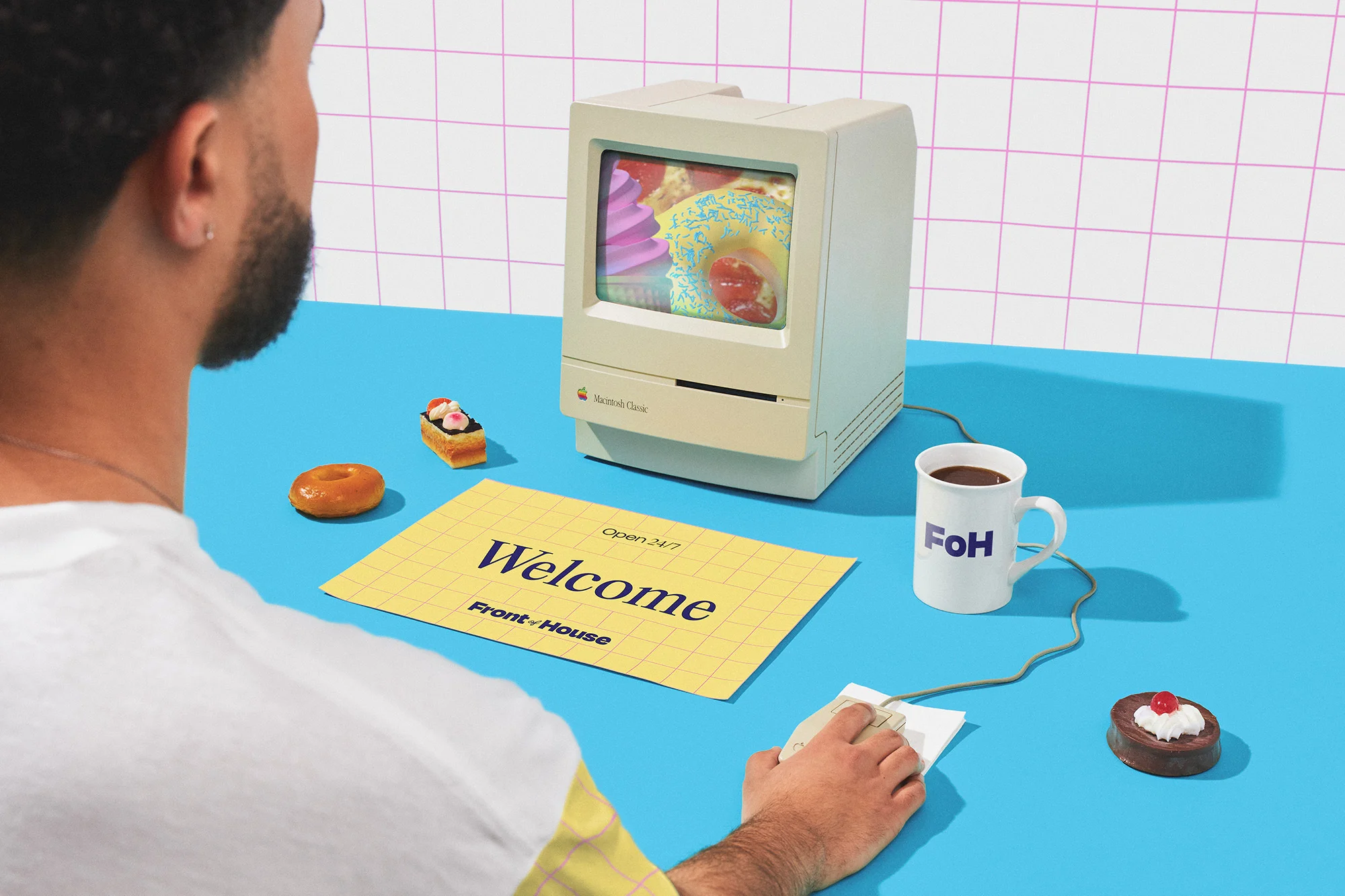 Person uses a retro Apple Macintosh computer with a dessert image on screen, alongside donuts and a cup on a blue table.
