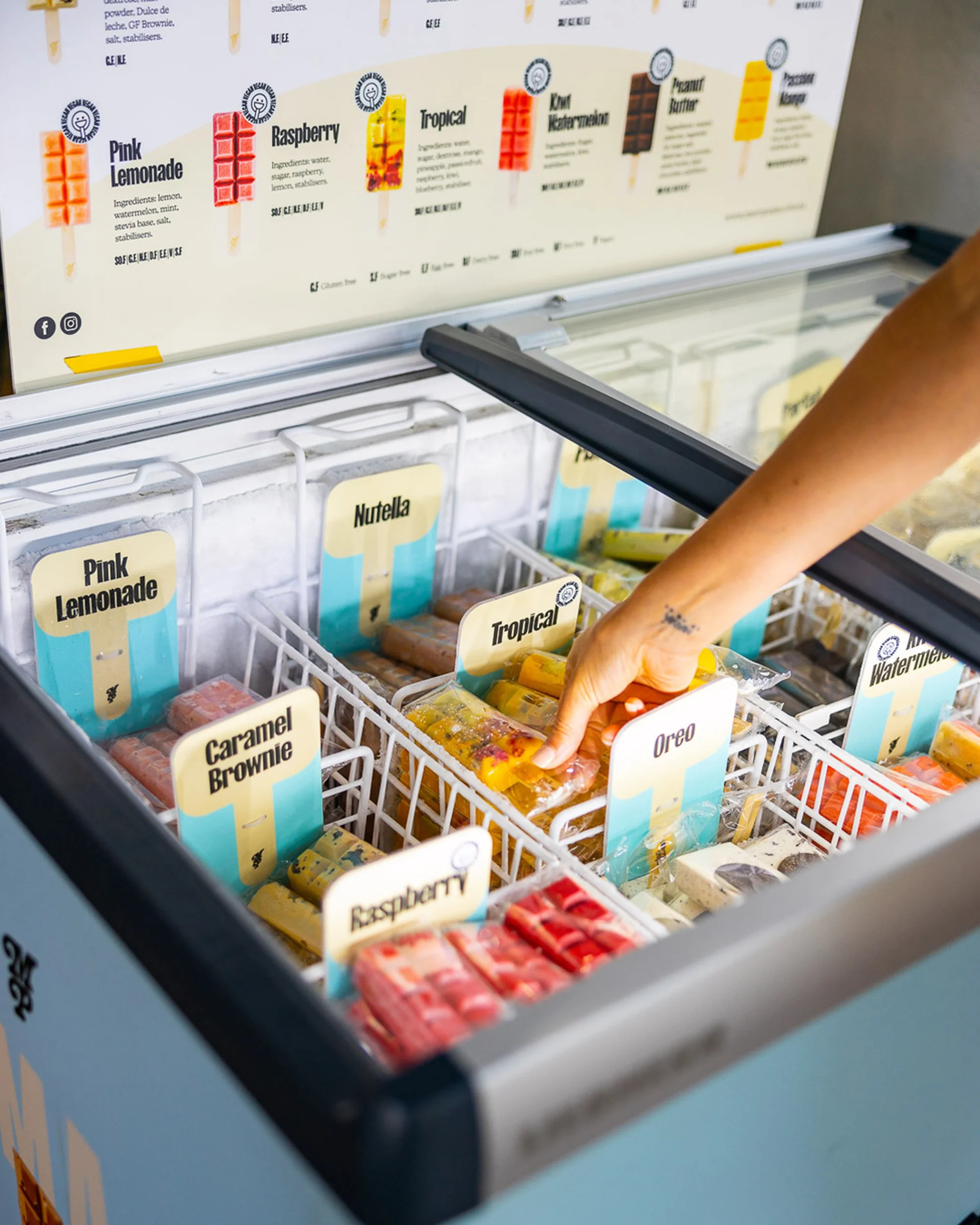 An arm reaches into a freezer containing various flavors of popsicles, including pink lemonade, caramel brownie, and tropical flavors.