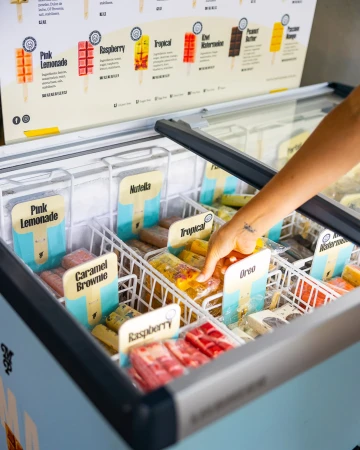 An arm reaches into a freezer containing various flavors of popsicles, including pink lemonade, caramel brownie, and tropical flavors.
