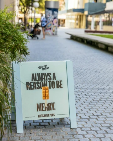 Outdoor shot of a Merry Pops ad sign on a sidewalk, featuring an ice pop image and text, with blurred figures in the background.