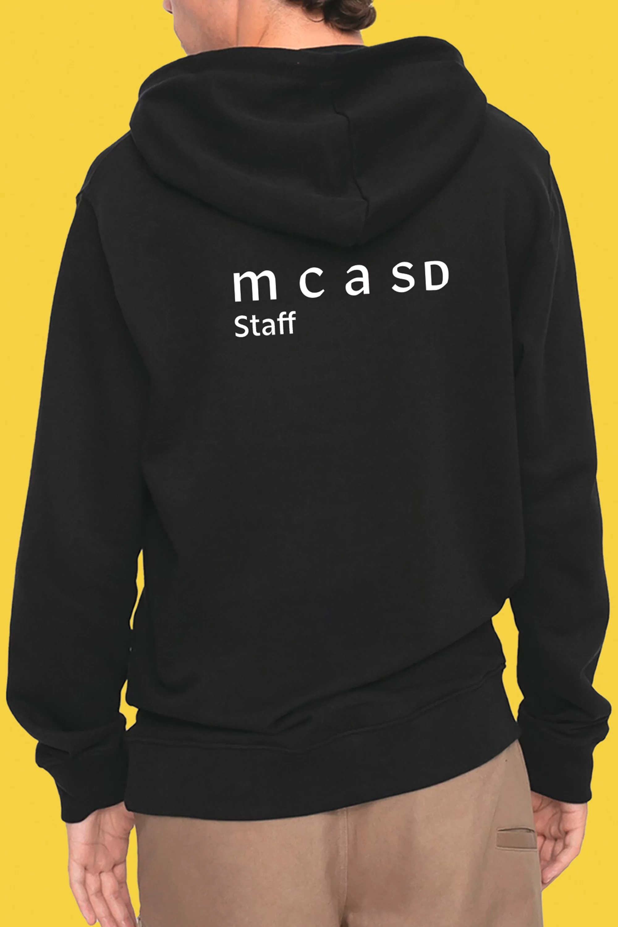 An individual wears a black hooded sweatshirt with 'mcaSD Staff' printed on the back, set against a vibrant yellow background.