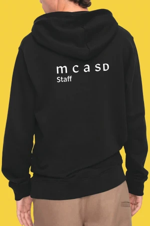 An individual wears a black hooded sweatshirt with 'mcaSD Staff' printed on the back, set against a vibrant yellow background.