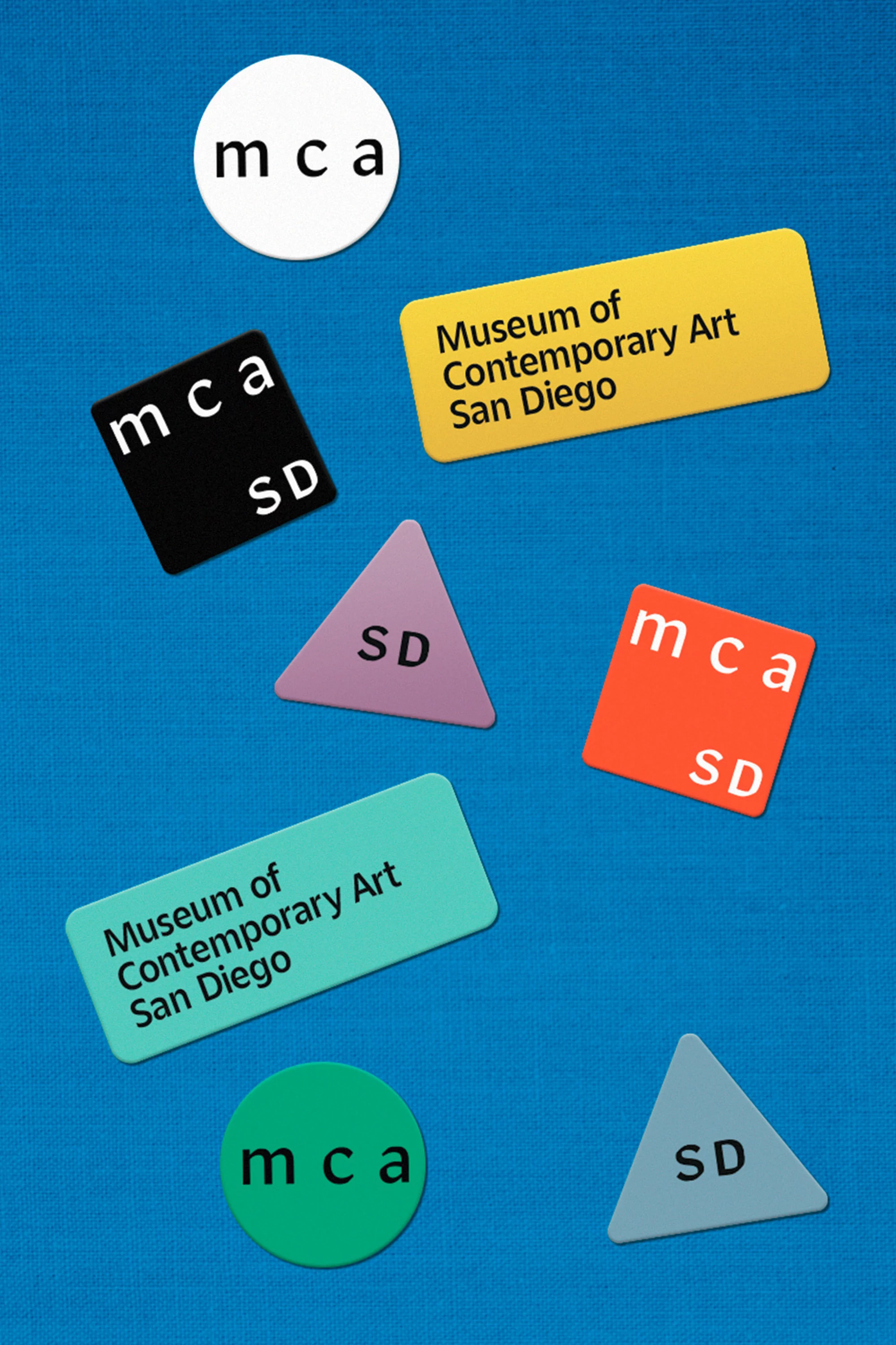 Floating MCA Museum of Contemporary Art San Diego logo badges of various colors and geometric shapes, displayed against a blue background.