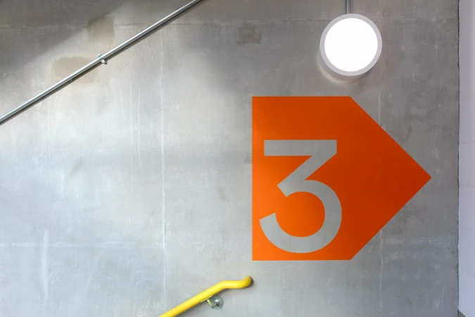 An orange arrow with the number 3 pointing right is stenciled on a gray concrete wall with a white light fixture and handrails.