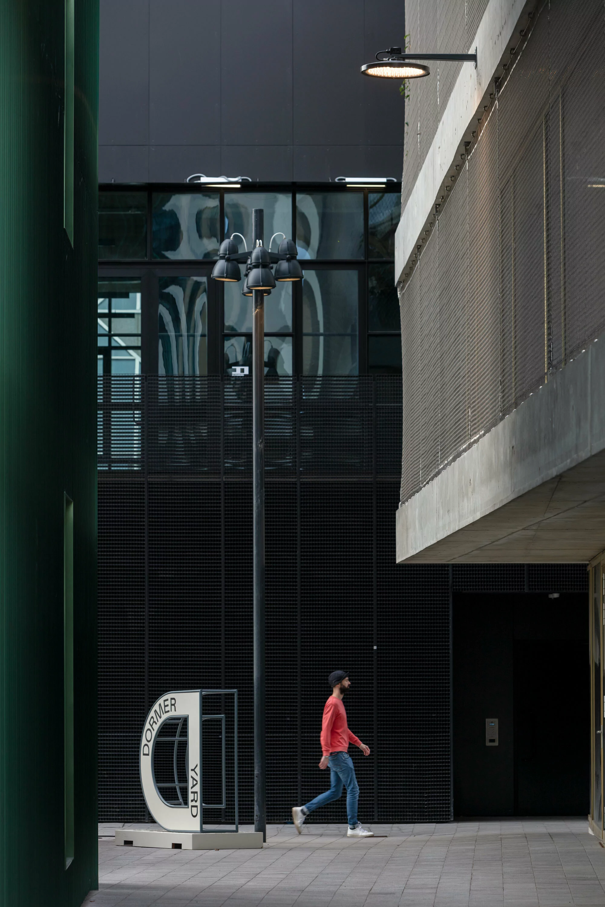 Adult male in red shirt walks past a Dormer Yard sign in an urban setting with modern architecture, street lights, and patterned sidewalks.
