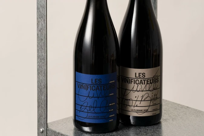 Two wine bottles with textured labels sit side-by-side on a metal shelf against a neutral background, showcasing beverage product photography.