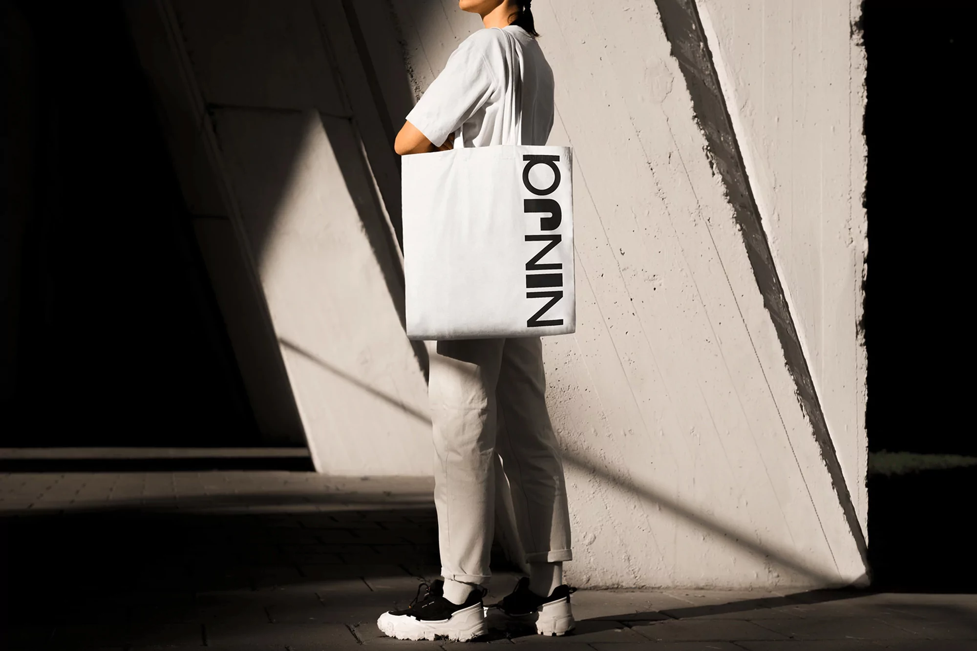 Woman in a white T-shirt and light pants holds a white tote bag with the word 'NINJA' printed on it, standing against a modern wall.