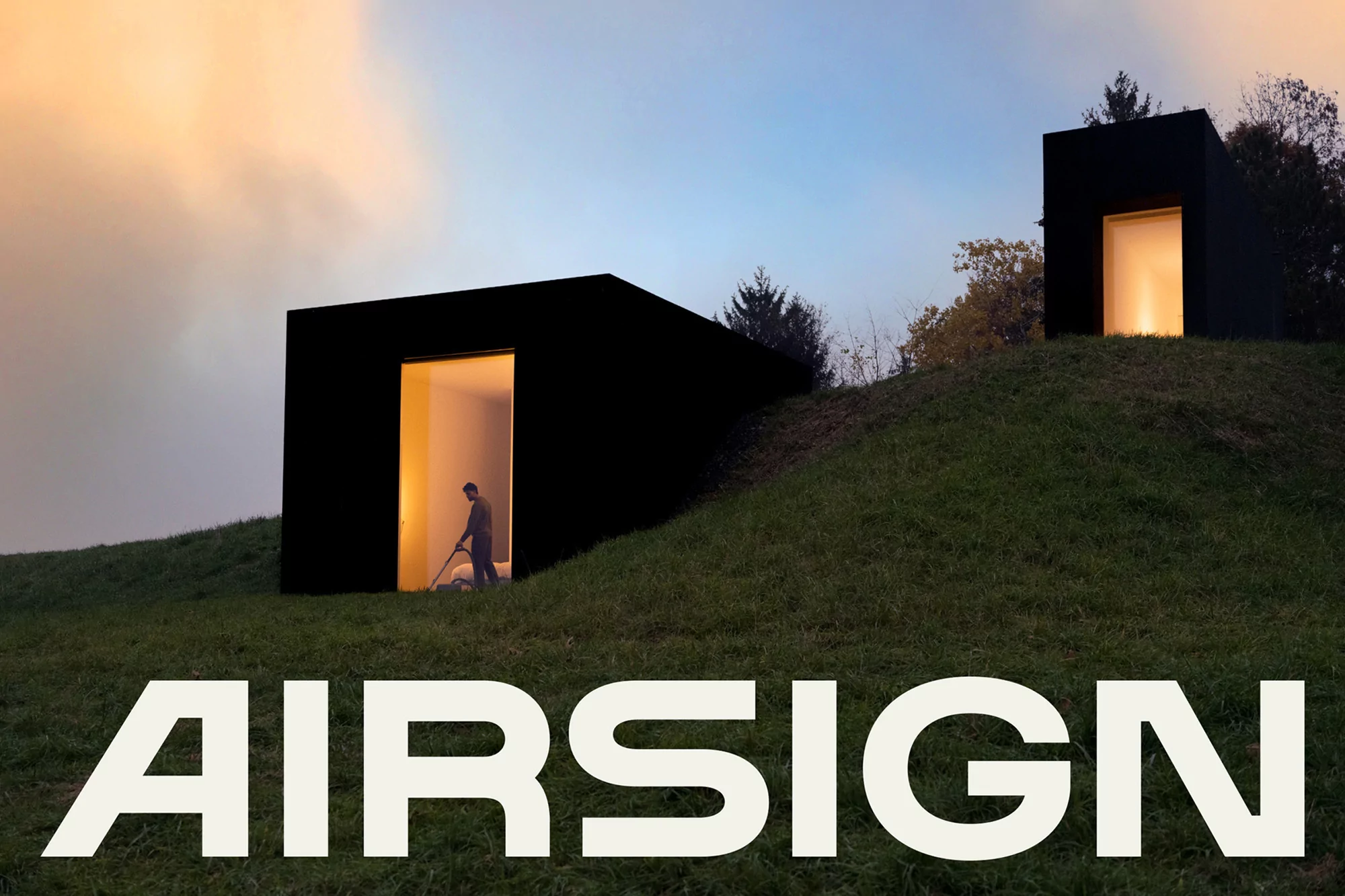 Modern black cube buildings on a grassy hill with a man vacuuming inside one, under a pastel sky, and the word AIRSIGN in bold letters.