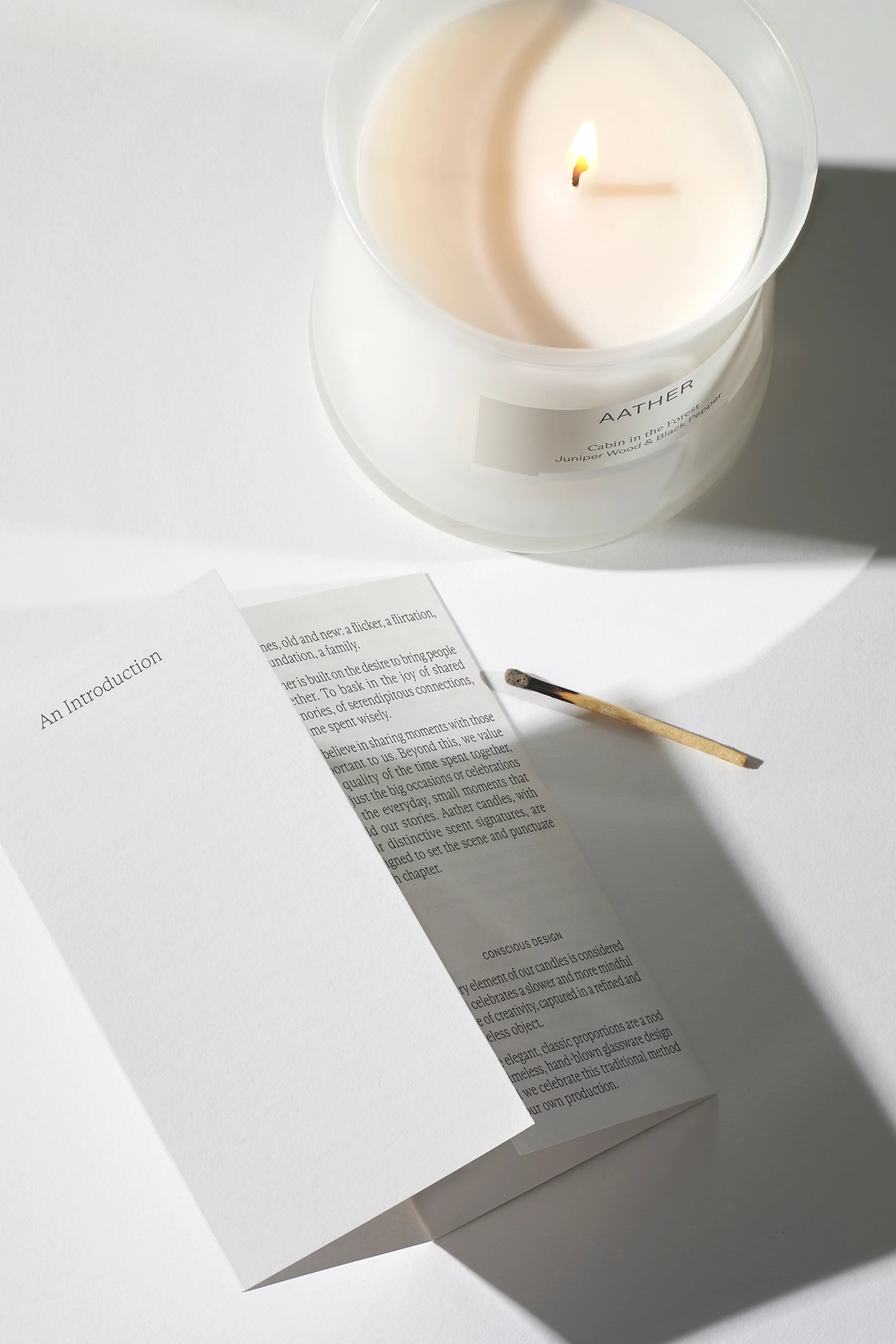 Still life featuring a lit candle in a glass jar, next to an open brochure, and a used matchstick on a white surface indoors.