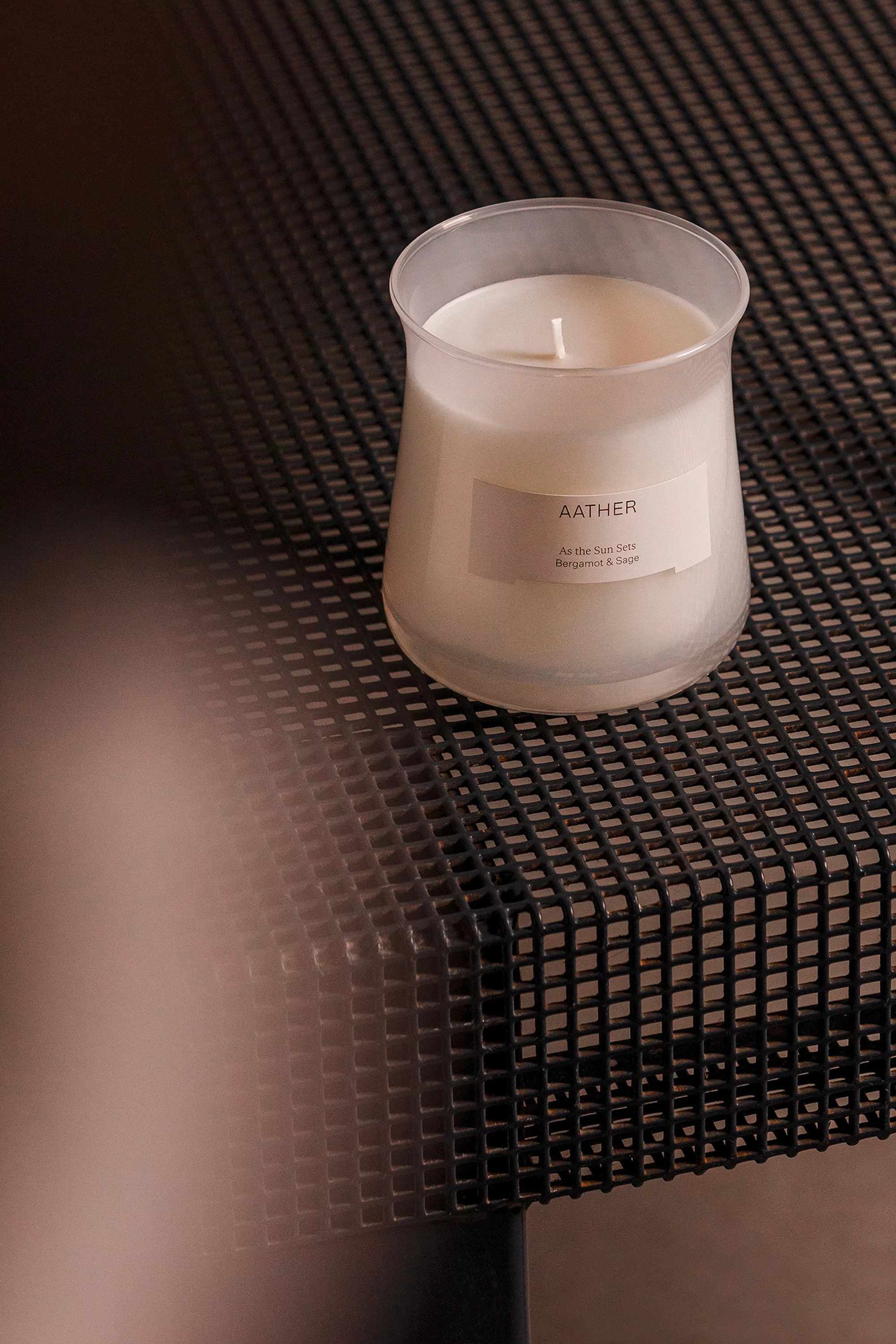 A white candle in a glass holder sits on a black wire-mesh table, creating a minimalist, modern, and aromatic ambiance in a room.